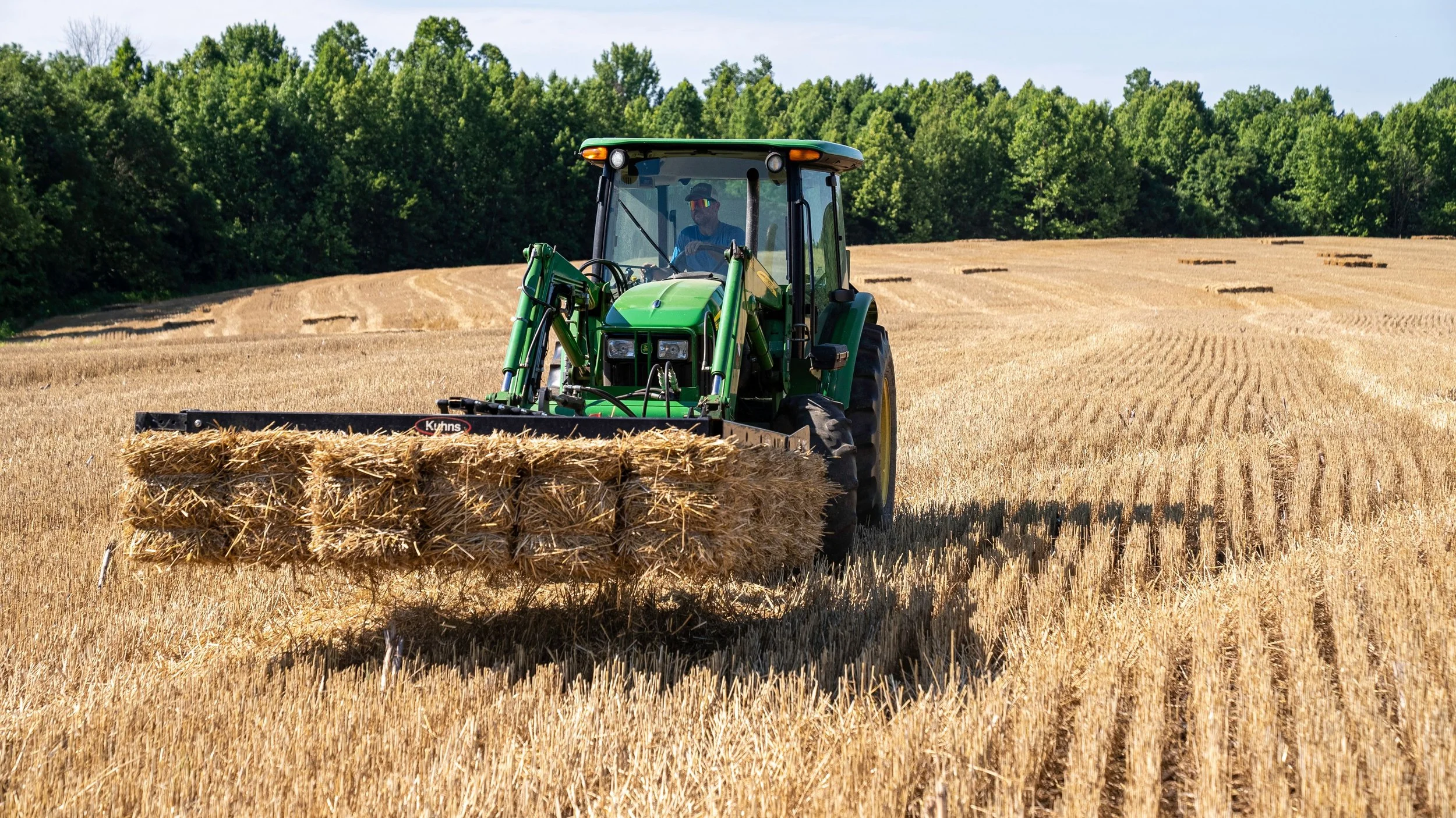 Wisconsin Farmers. Dairy Farm. Agriculture. US Farm Bill. A man driving a green tractor with a front attachment in a golden wheat field, with trees in the background on a clear day.
