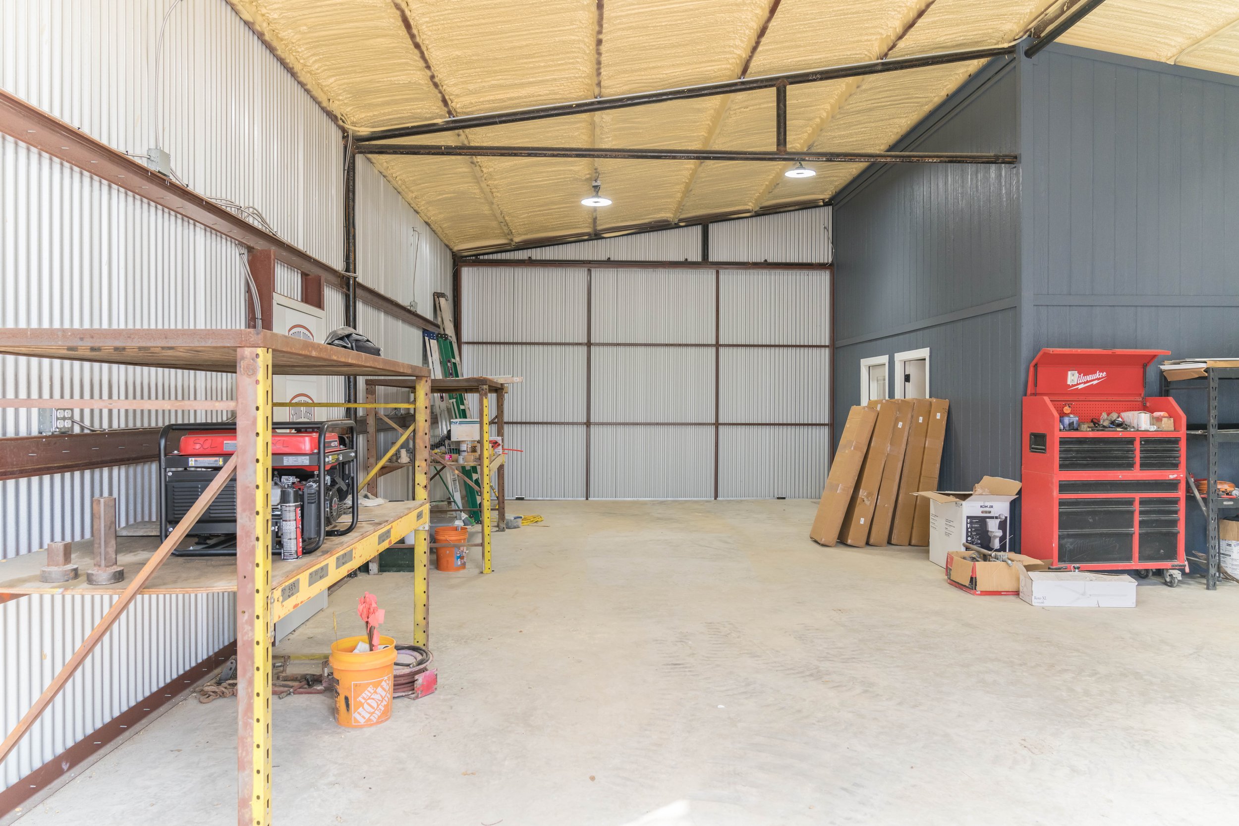 Empty garage with metal siding walls, a concrete floor, and a partially finished ceiling. Shelves and tools are on the left side, with a ladder and boxes on the right. There is a large red toolbox and some wood leaning against the wall.