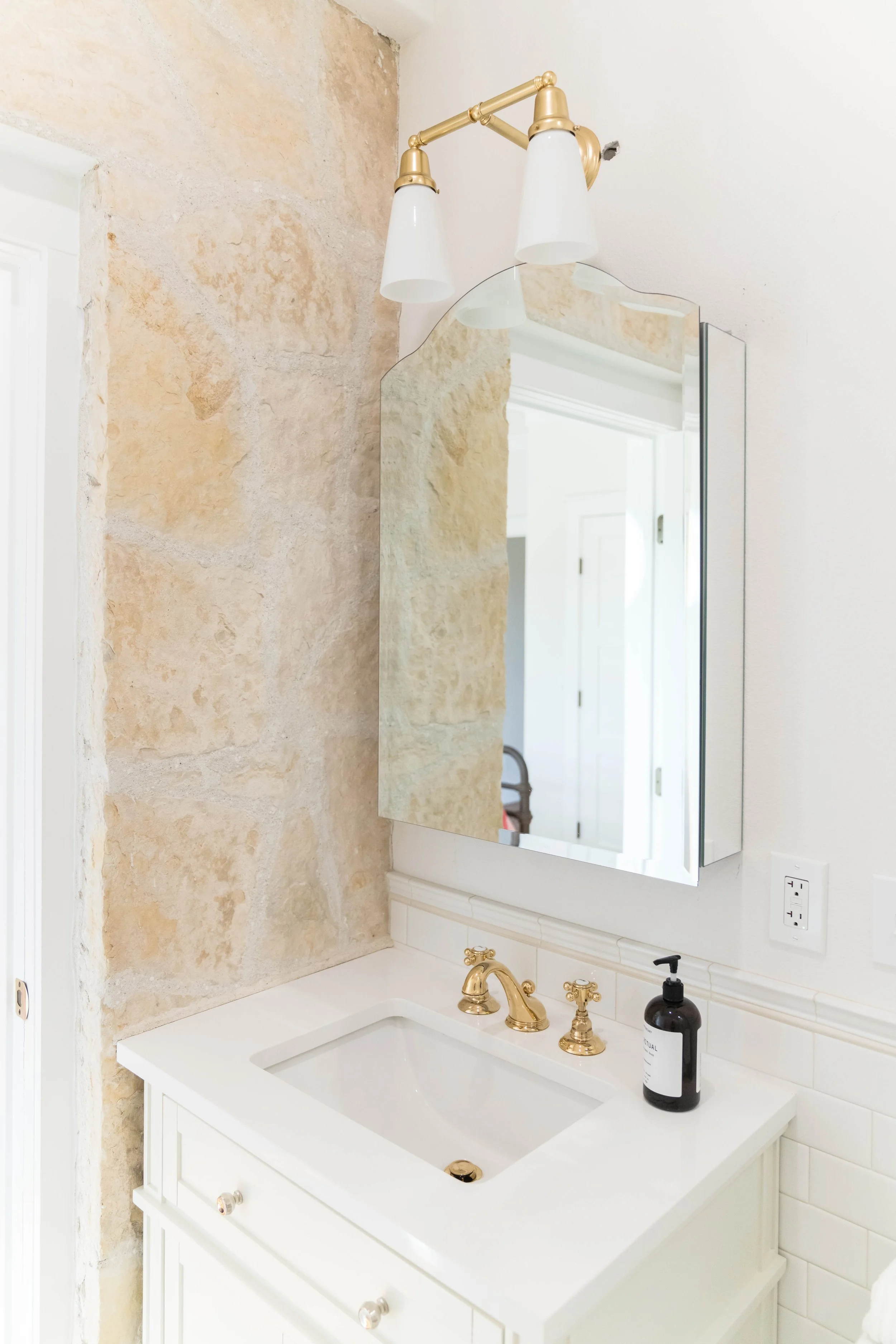 Bathroom vanity with a white countertop, gold fixtures, black soap dispenser, and a mirror cabinet. Wall has a mix of white paint and natural stone finish. Overhead light fixture with three white shades and gold accents.