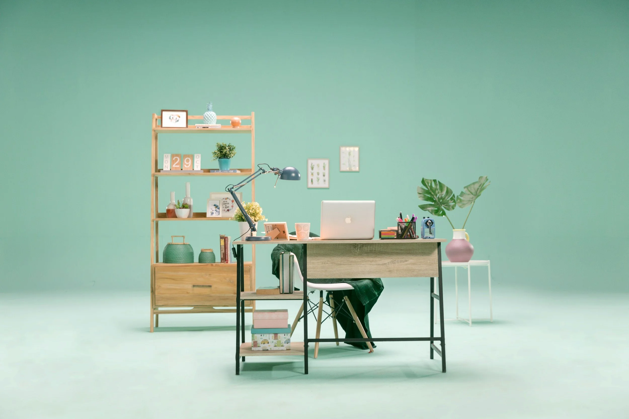 An office workspace with a mint green wall and a desk holding a laptop, notebook, pen holder, and plant. Behind the desk is a wooden bookshelf with decorative items, books, and picture frames. There is a small side table with a pink and white vase and a large leafy plant. The floor is mint green.