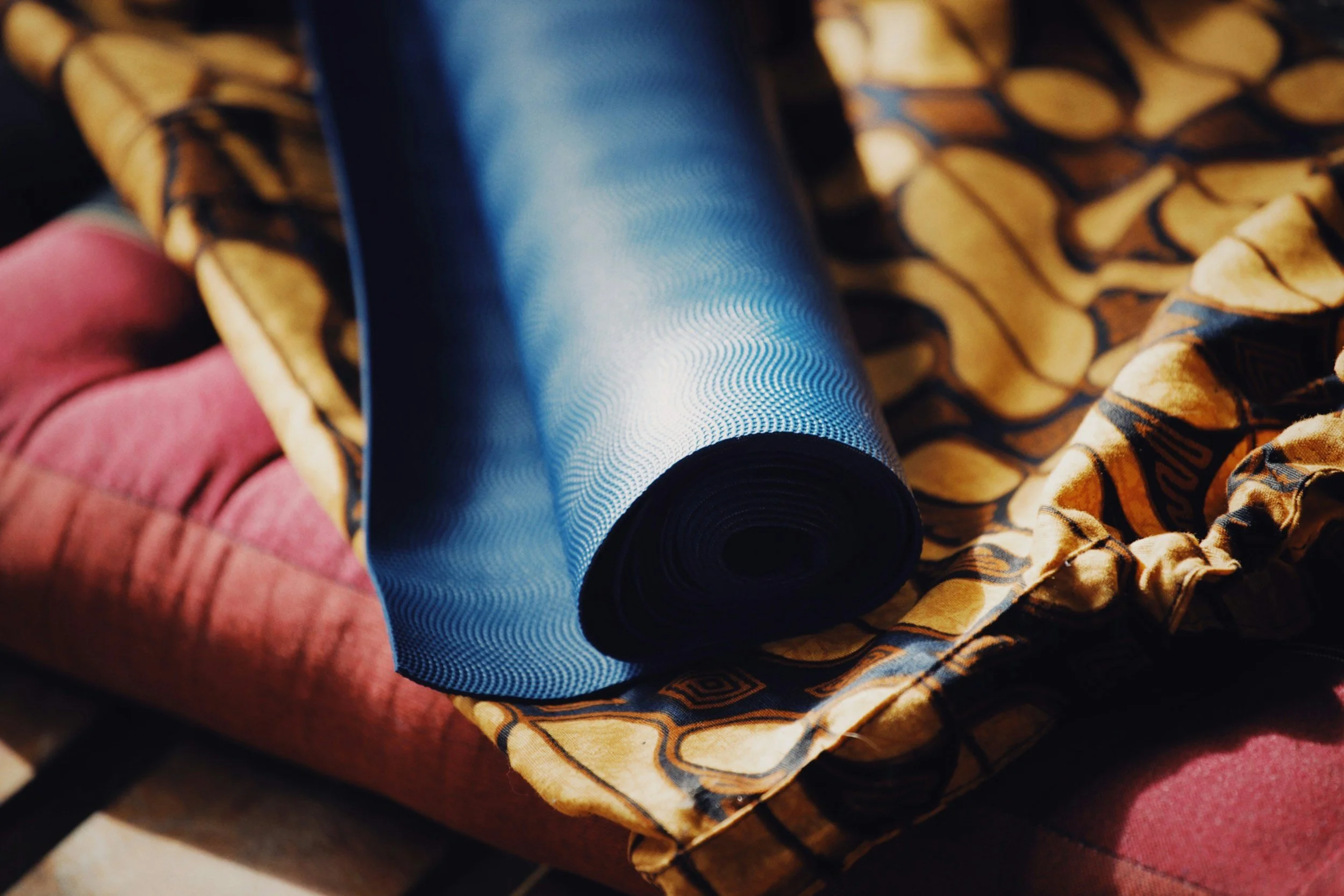 Close-up of fabric rolls, including a patterned yellow and black fabric, a blue textured fabric, and a pink fabric, resting on a reddish surface.