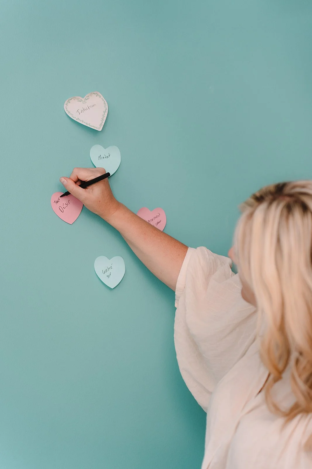 Émilie Evans writing keywords onto heart-shaped sticky notes on a turquoise wall, illustrating goal setting and personal transformation.