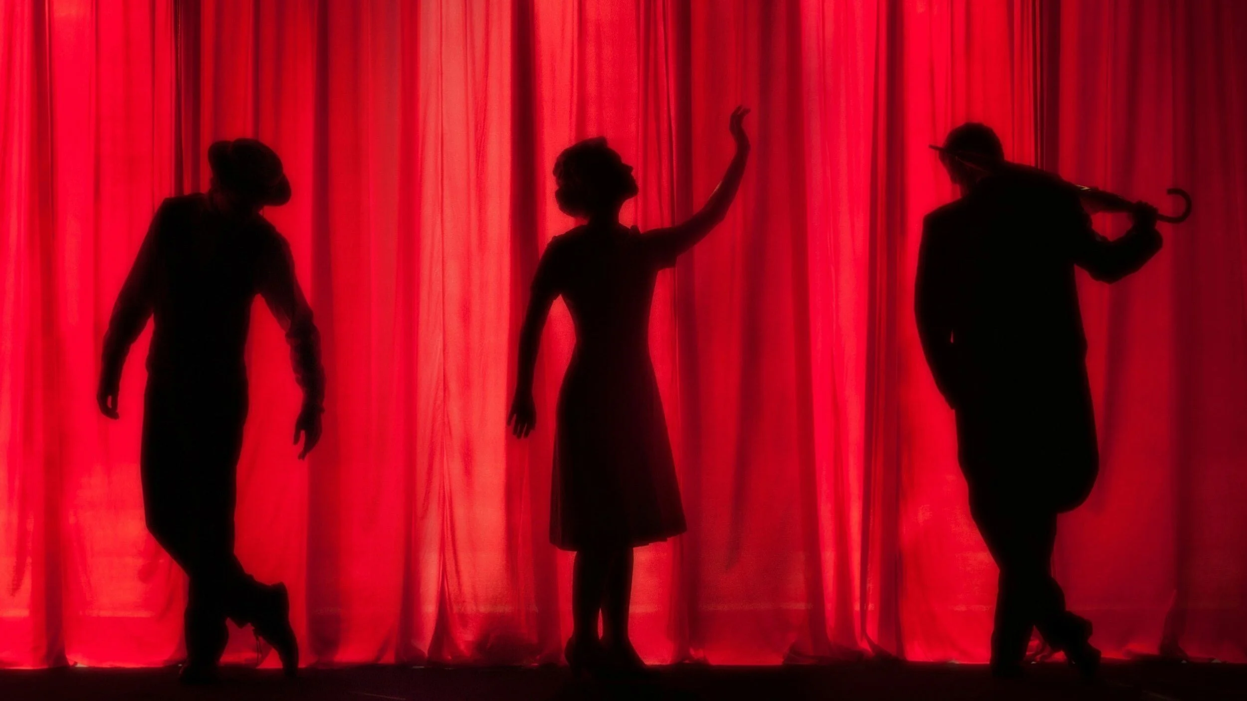 Three silhouettes strike theatrical poses at the stage floor’s front edge, framed by red curtains and dramatic architecture.