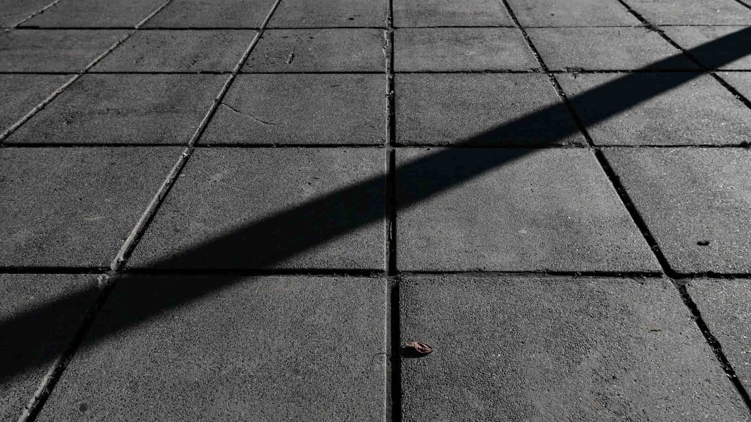 Shadow stretched diagonally across sidewalk-concrete walkway paving in sunlight, with a small brown leaf on the ground.