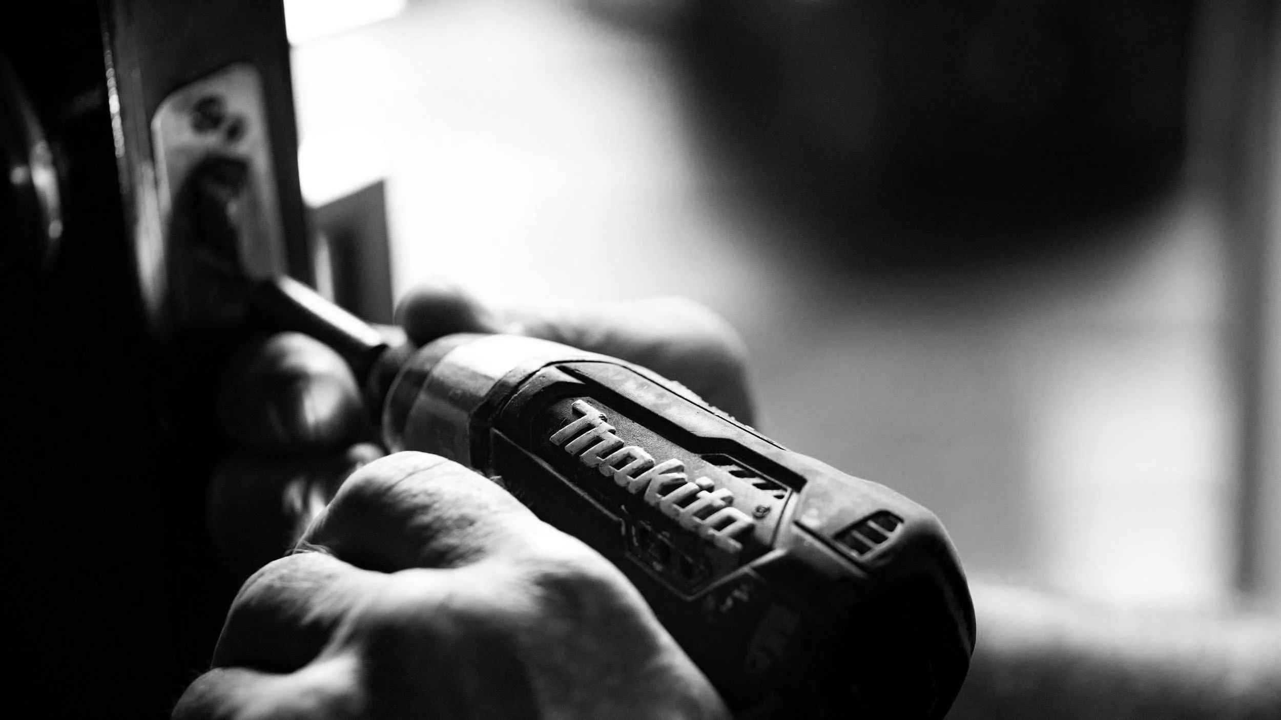 Close-up of hands using a Makita power drill to secure surface-anchored counterflashing or a metal plate on a door, in black and white.