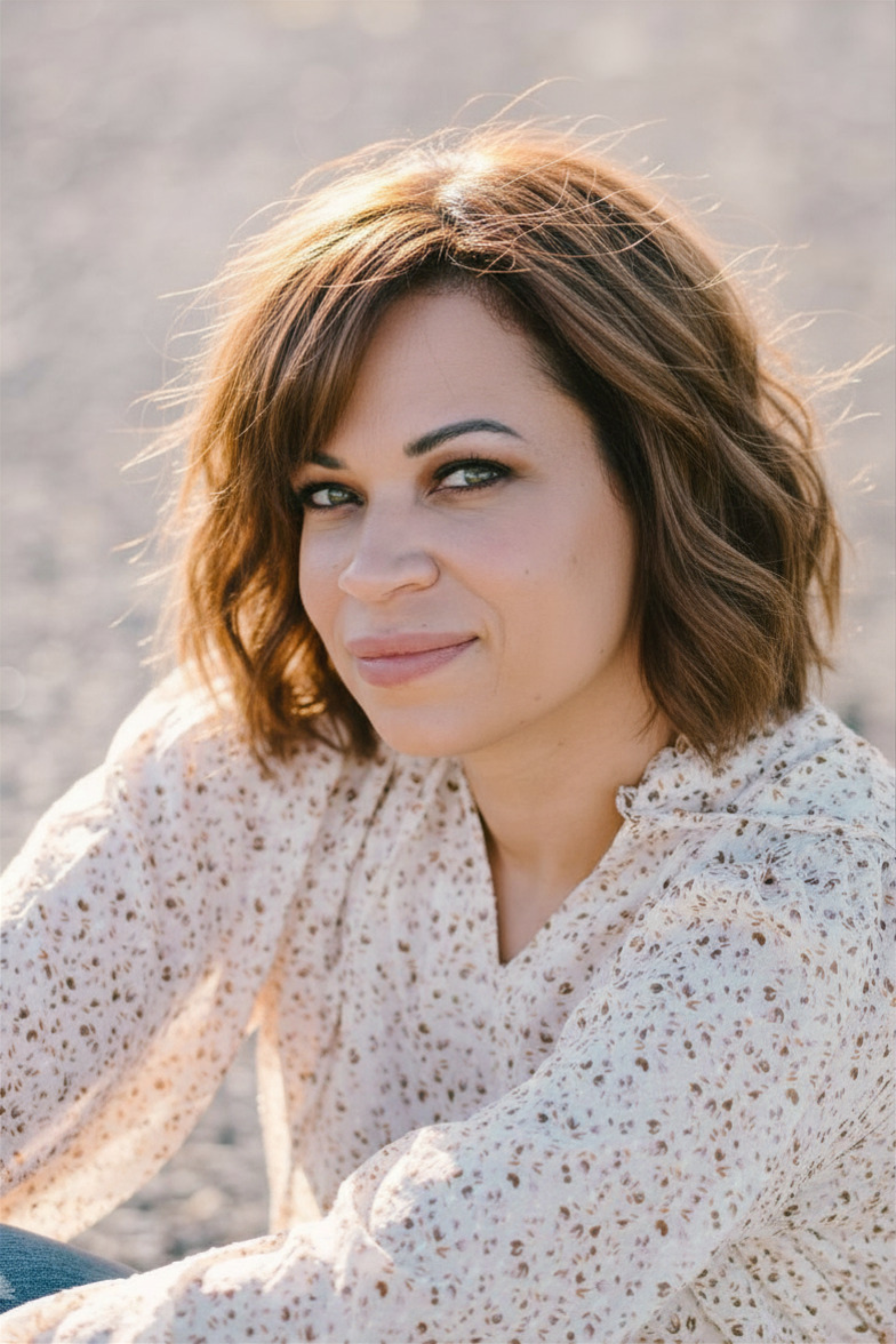 A woman with shoulder-length brown hair and light makeup, wearing a cream-colored, long-sleeved blouse with small brown patterns, sitting outdoors with a blurred background, looking at the camera with a slight smile.