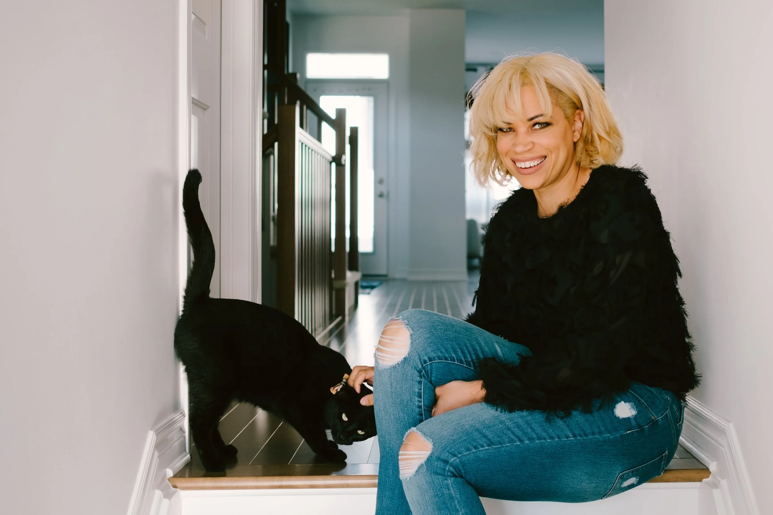 A woman with blonde hair, wearing a black fuzzy sweater and ripped jeans, is sitting on the floor smiling at the camera. She is interacting with a black cat that is sniffing her hand in a home corridor with a staircase and a door in the background.