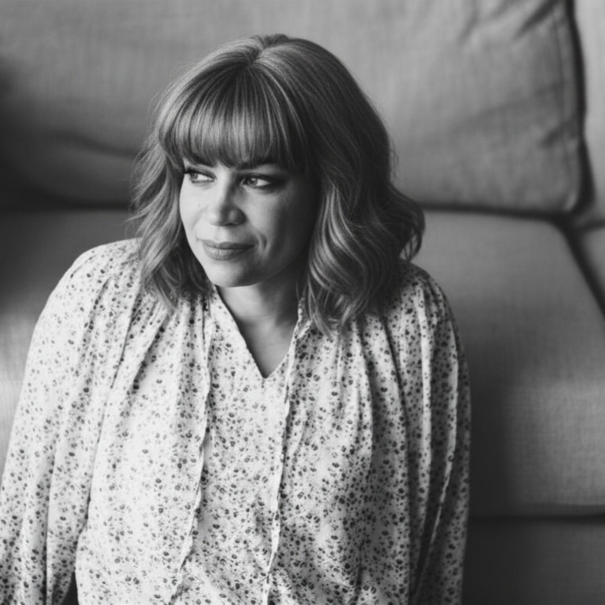 Black and white portrait of a woman with shoulder-length wavy hair, wearing a patterned blouse, sitting against a cushioned background, looking to her left.