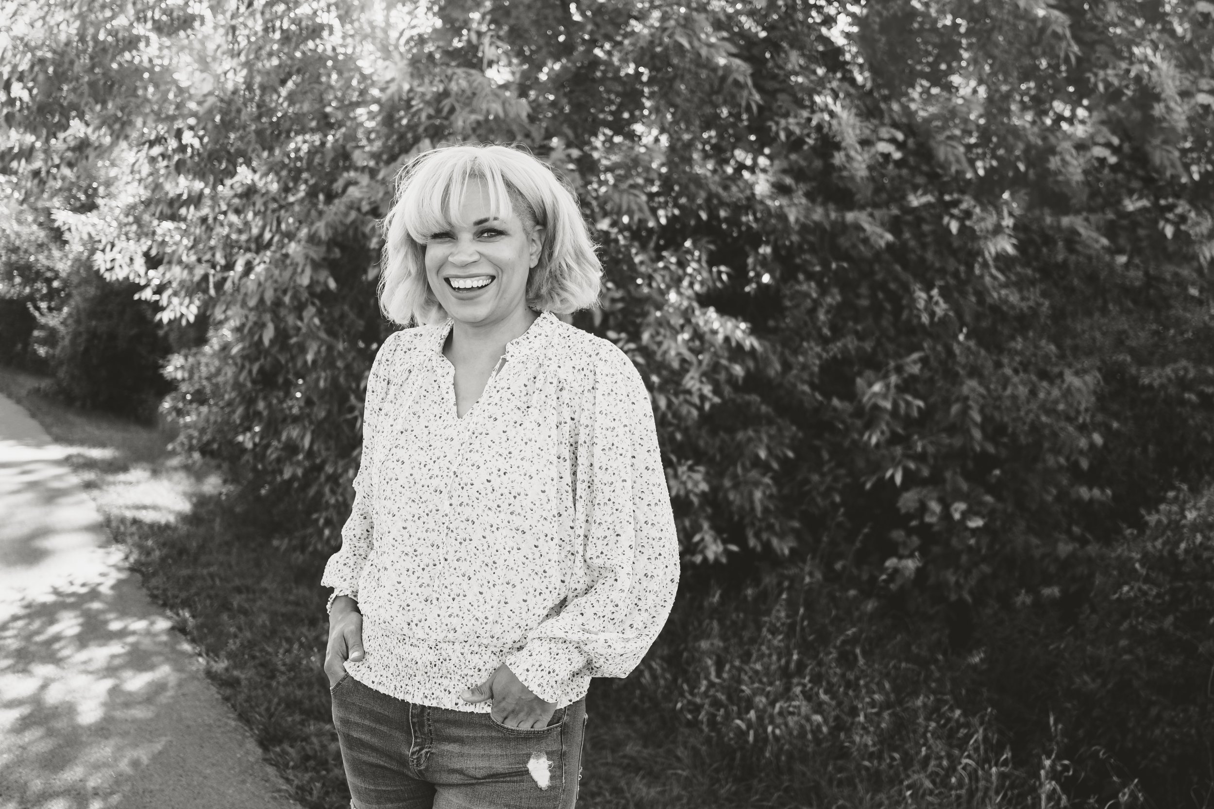 A woman with blonde hair in a bob style, winking and smiling, standing outdoors in front of bushes and trees, wearing a patterned long sleeve shirt and ripped jeans.