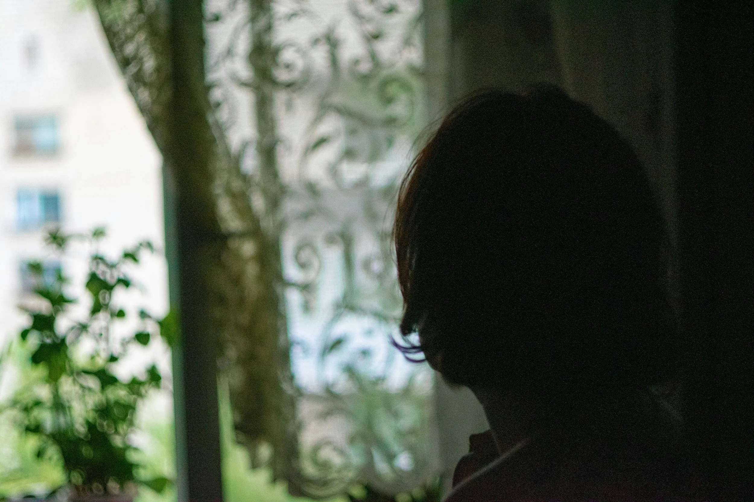 A woman inside a dark room looking outside through an out-of-focus window