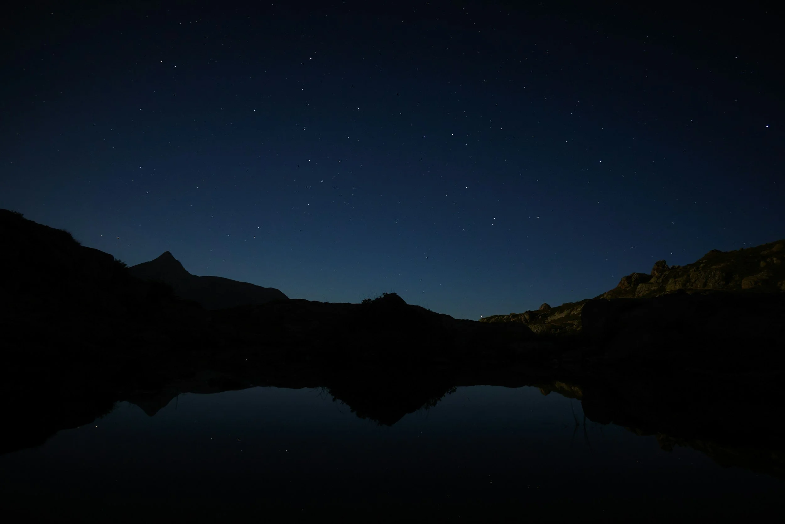 Nighttime landscape with a clear starry sky, mountain silhouettes, and a reflection of the mountains and stars on a calm body of water.