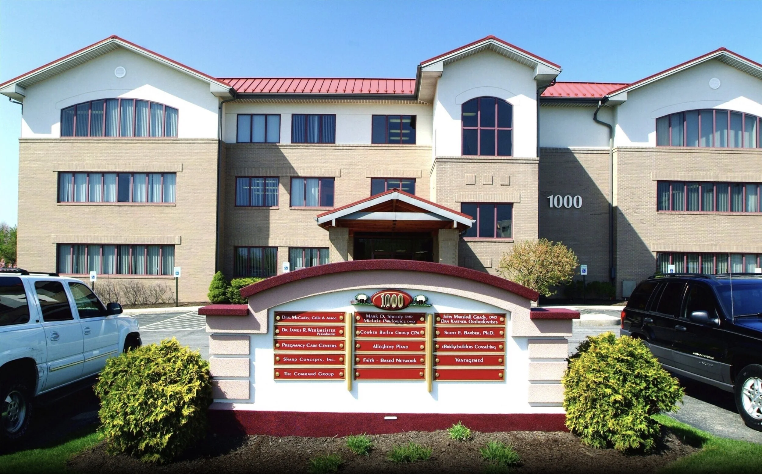 Exterior view of a multi-story office building with beige brick walls, red roof, and numerous windows. In front, there is a sign with the address 1000 and a list of business names. Several cars are parked nearby, and there are small bushes and landscaped plants around the sign.