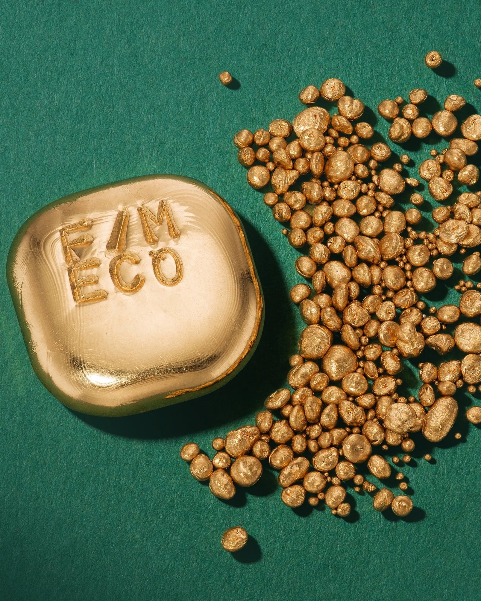 Gold-colored soap with the words 'FEM ECO' engraved on top, and a pile of small gold-colored pebbles scattered beside it, all on a green surface.