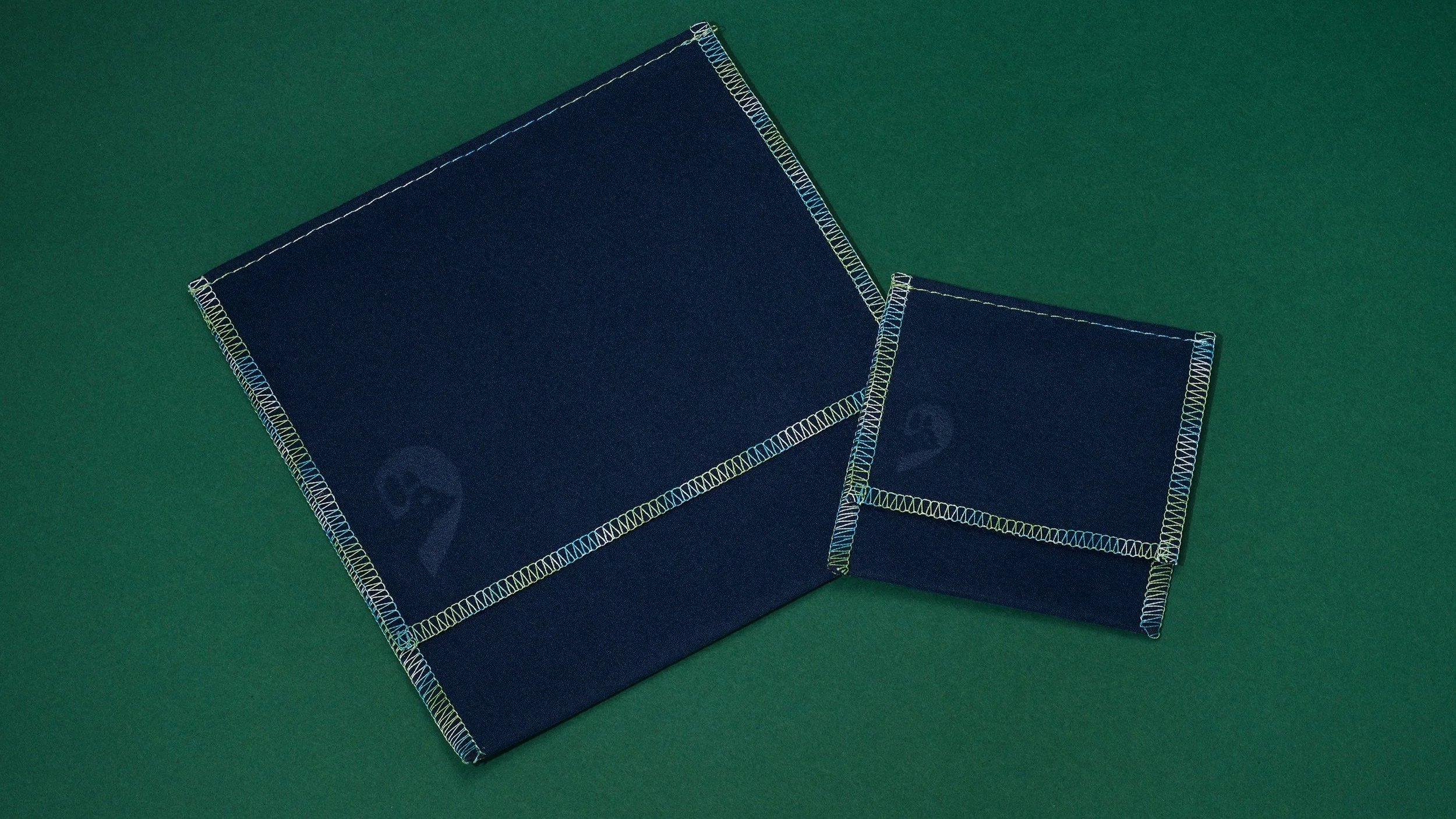 Two dark blue fabric-covered boxes with colorful stitched edges, one larger and one smaller, placed on a green surface.