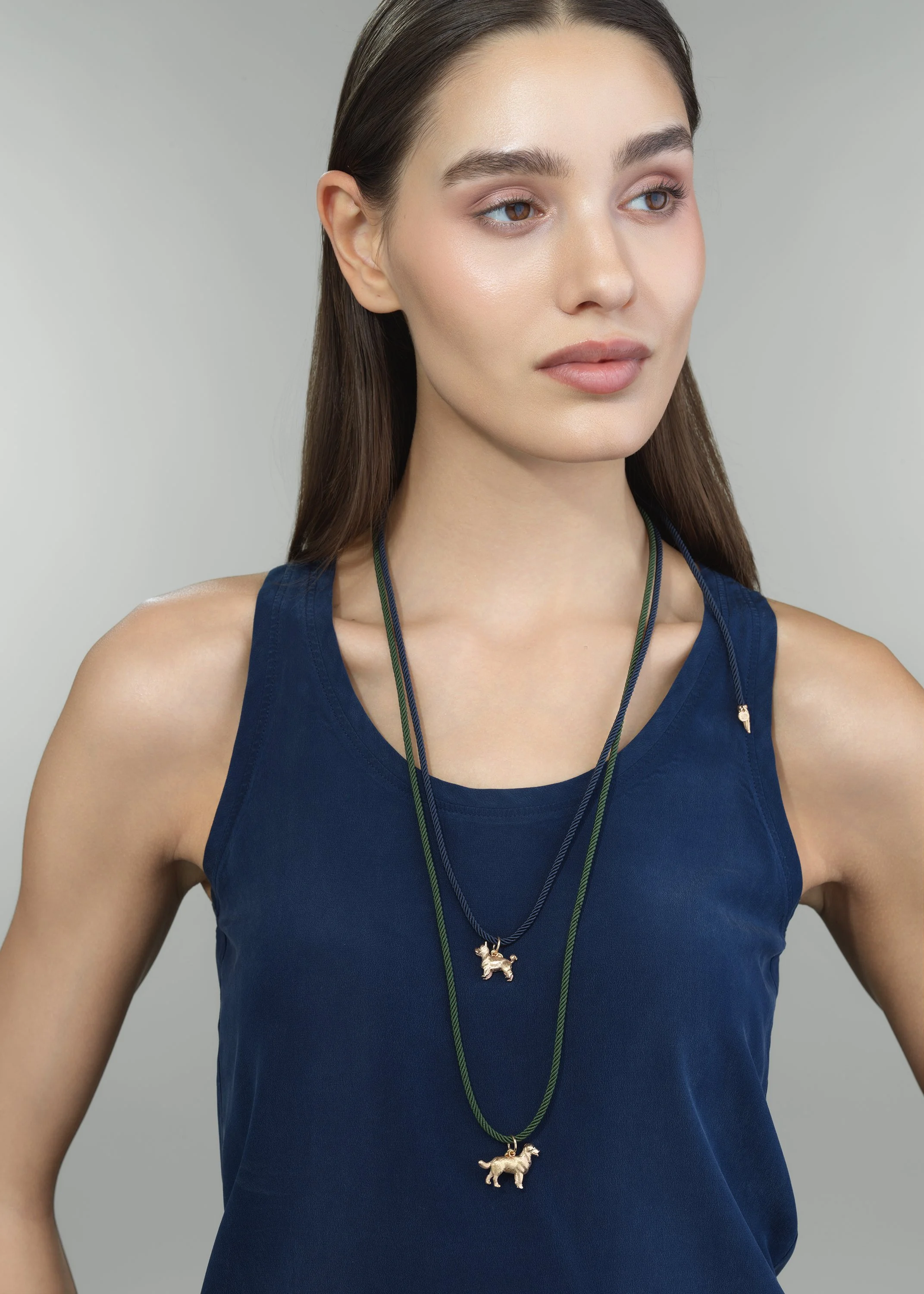 A young woman with long brown hair, wearing a navy blue sleeveless top and two layered necklaces with dog-shaped charms, standing against a plain light gray background.