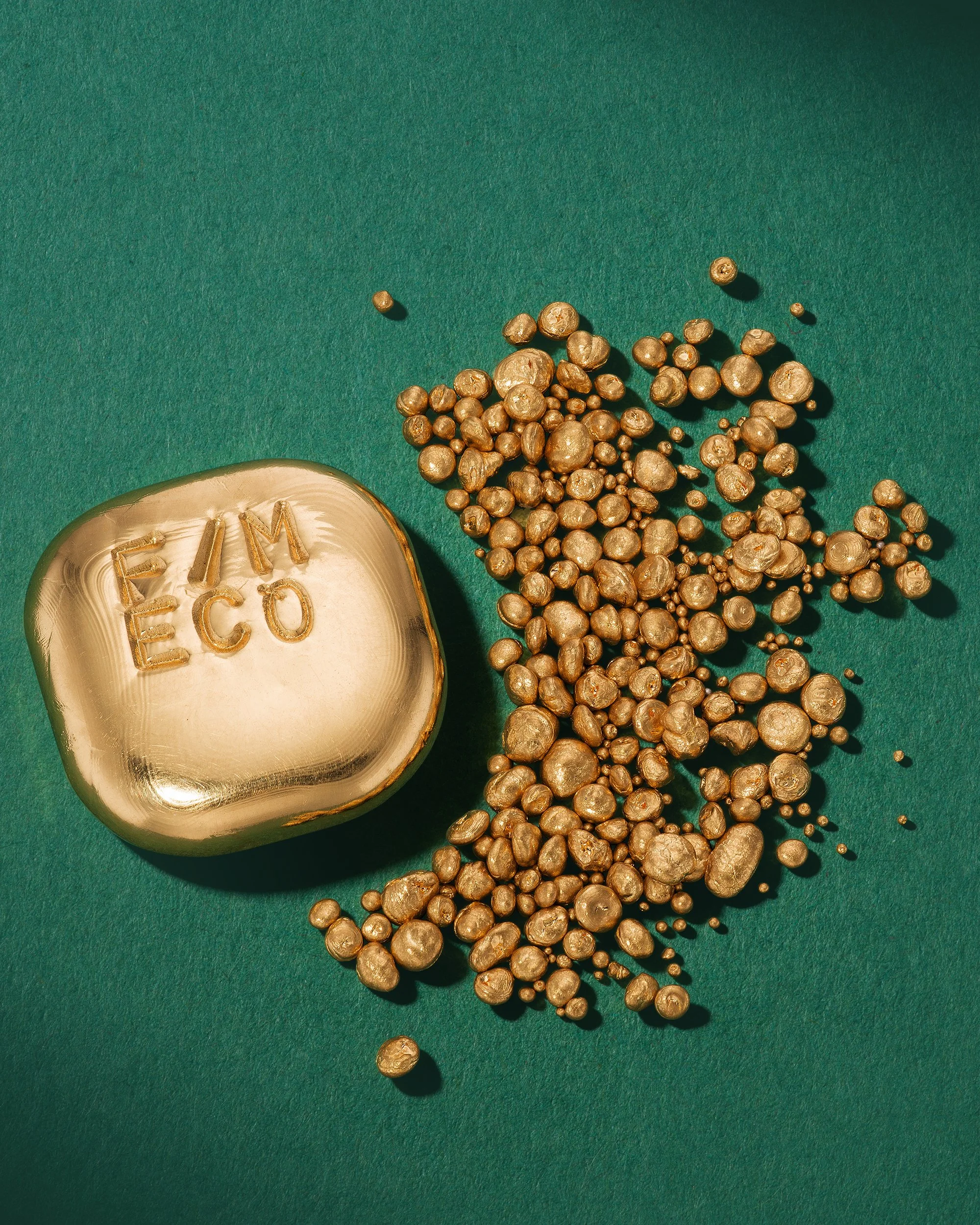 Gold soap with 'EIM ECO' engraved on it, surrounded by scattered gold-colored beads on a green background.