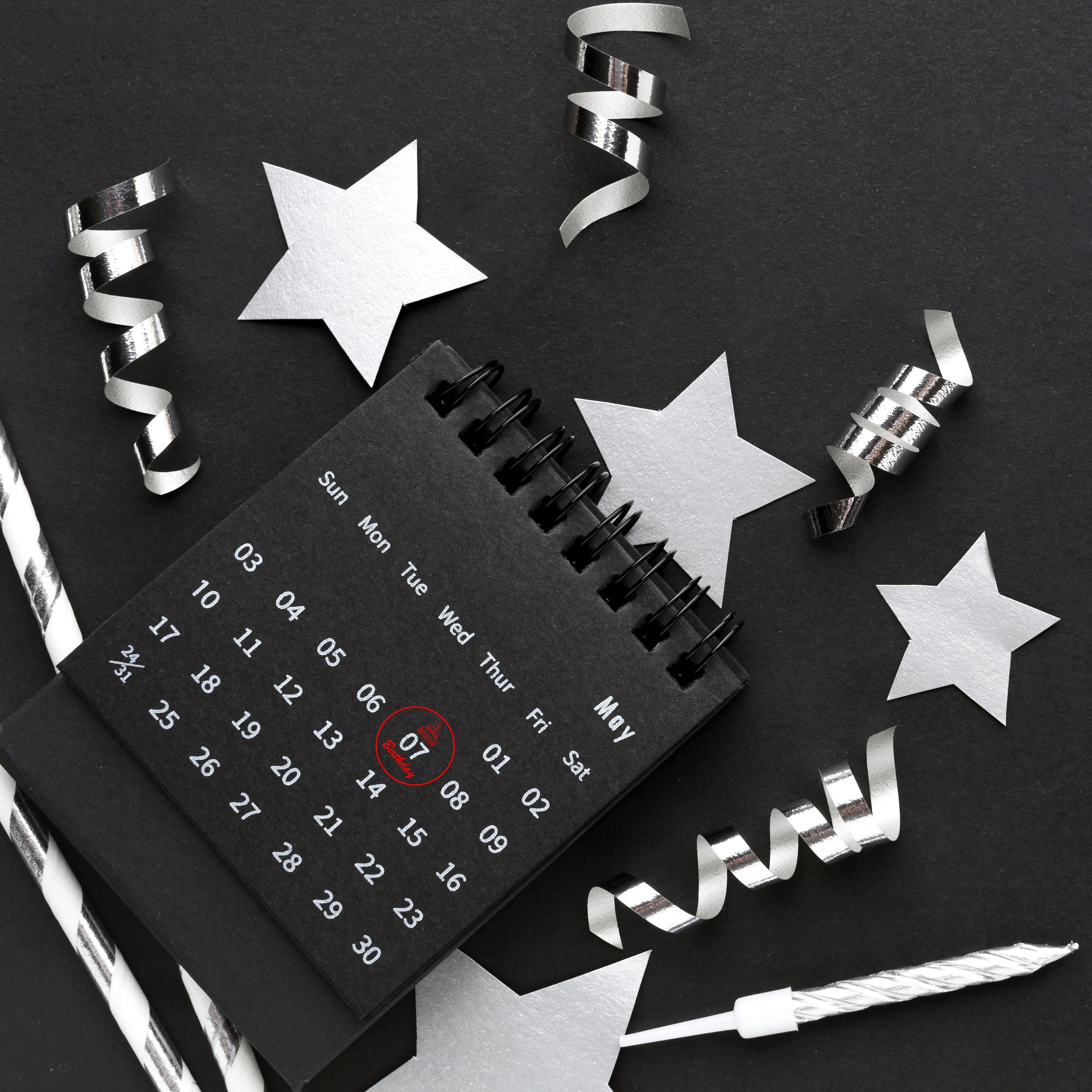 A black desktop calendar marked with May 7th, surrounded by silver and white party streamers, paper stars, and a white and silver party horn.
