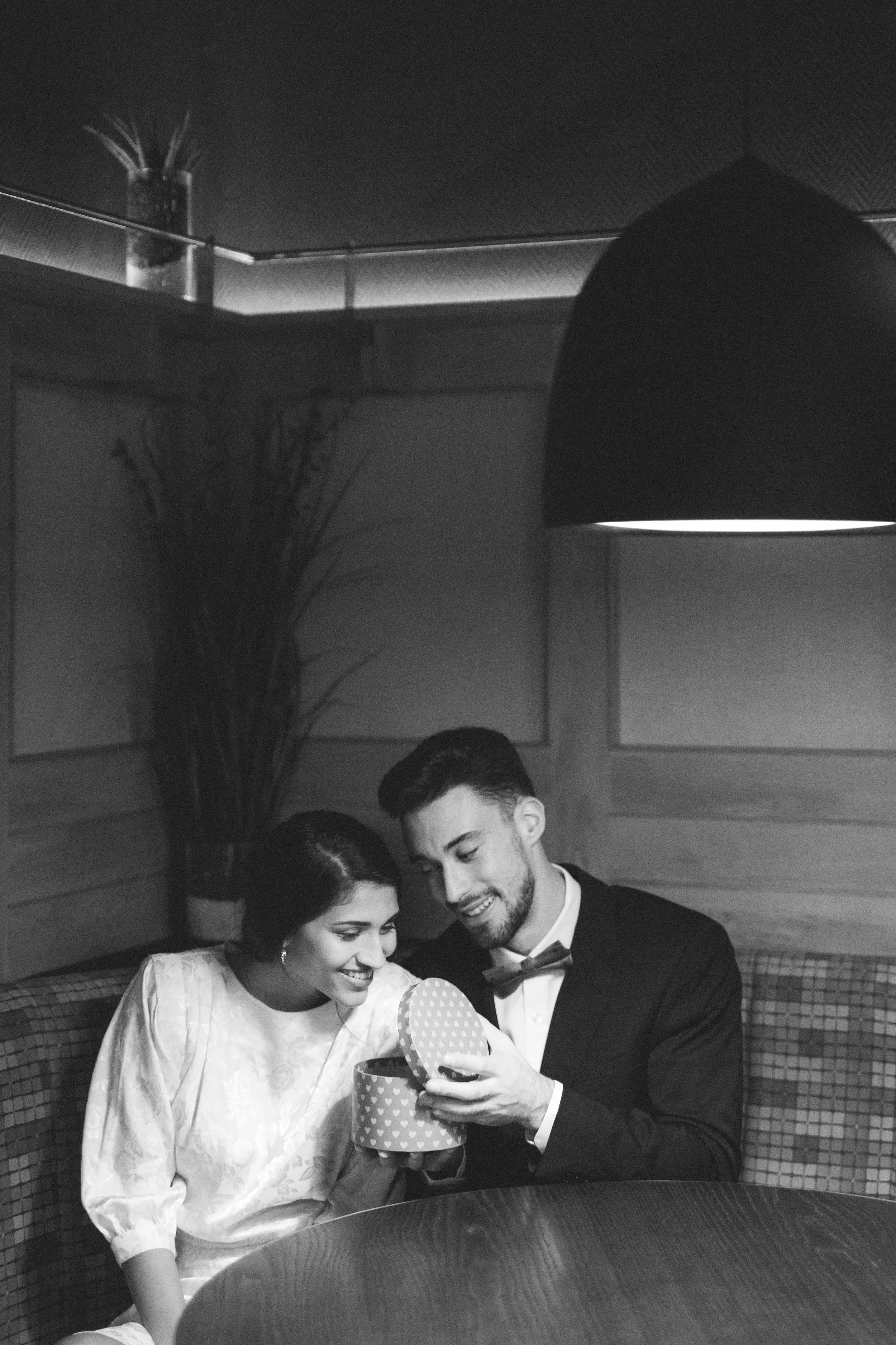 A black and white photo of a smiling couple sitting at a table, woman in a white dress and man in a tuxedo, both looking into a polka-dotted gift box held by the man.