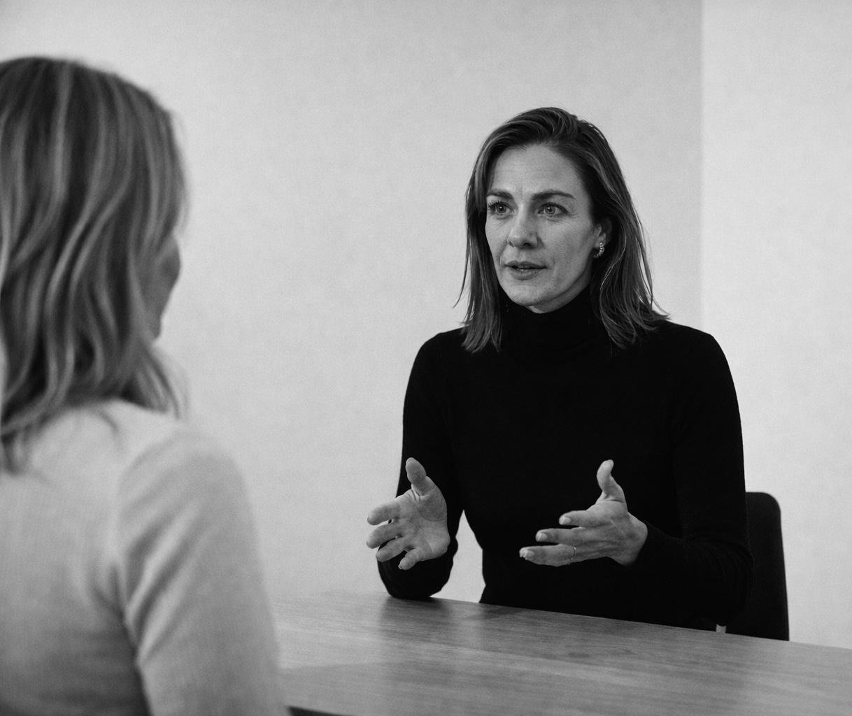 A woman with shoulder-length hair and a black turtleneck talking to another woman with blonde hair, blurred in the foreground, while seated at a table in a plain room.
