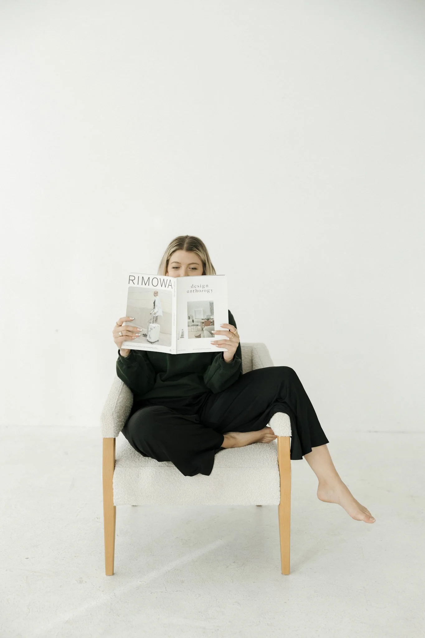 A woman sitting on a modern, light-colored chair with wooden legs, reading a magazine, with one foot on the ground and the other foot tucked under her.