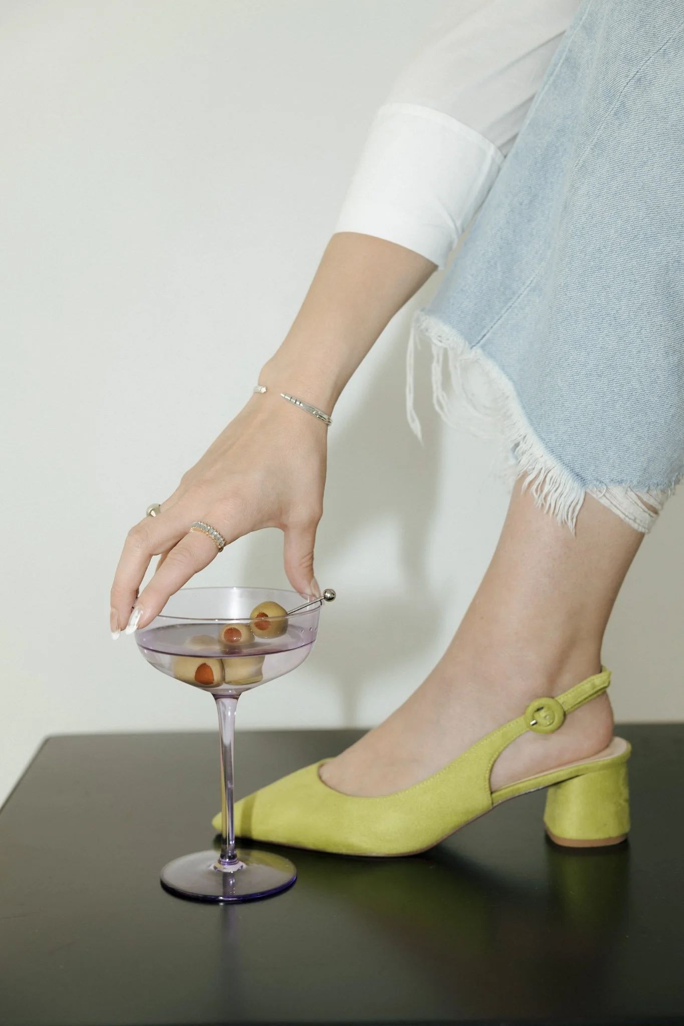 Close-up of a woman wearing yellow heels and distressed denim shorts, reaching to pick up a purple cocktail glass with olives, on a black surface.