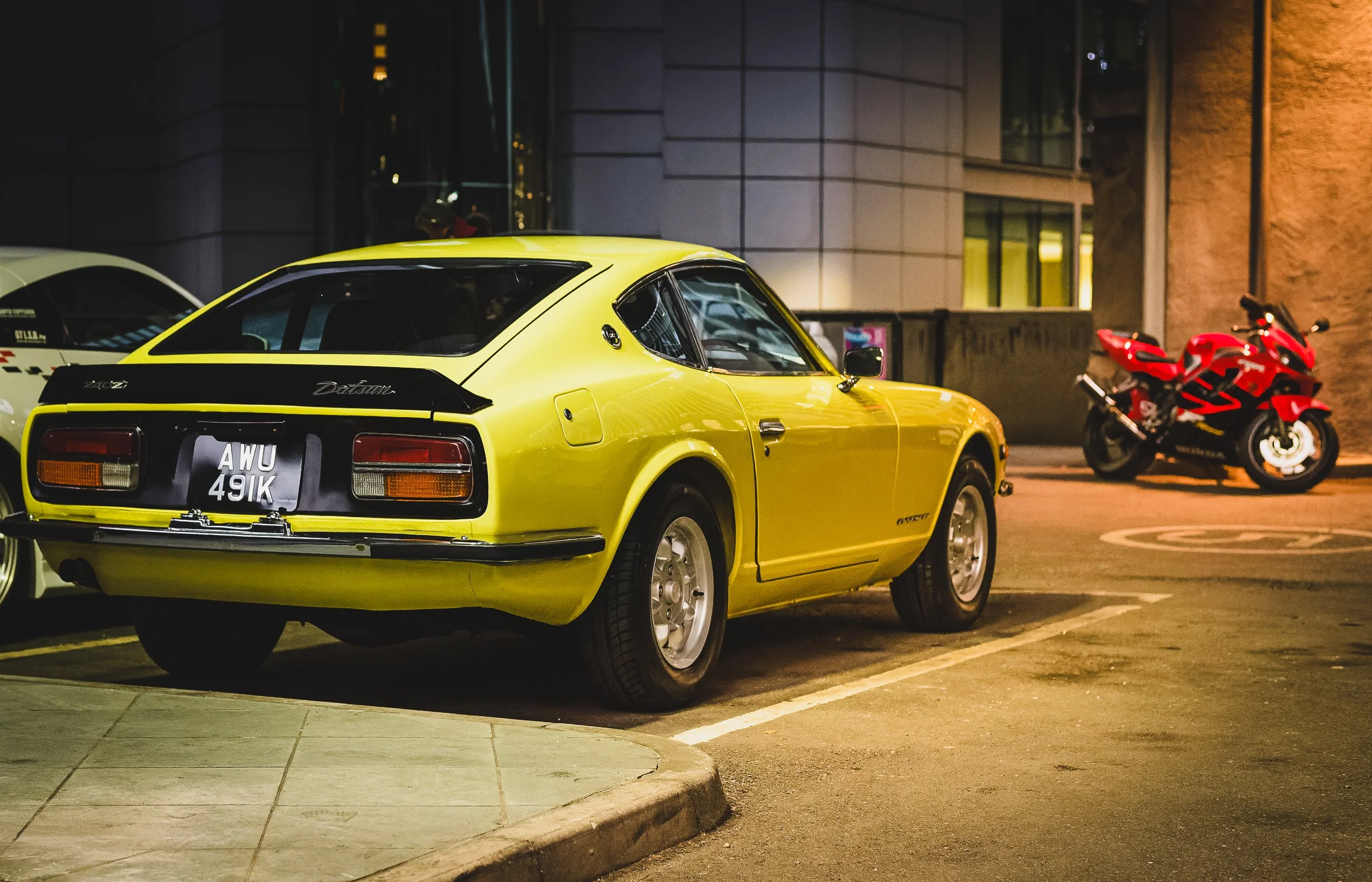 A yellow vintage Datsun 240Z sports car parked in a parking lot at night, with a red motorcycle in the background.