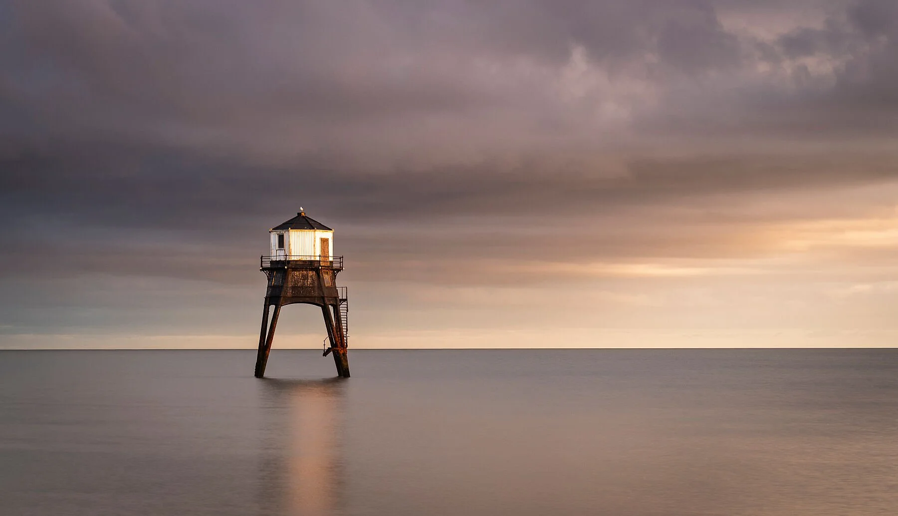 A solitary lighthouse on stilts in calm water, with a cloudy sky and soft, warm sunlight in the background.