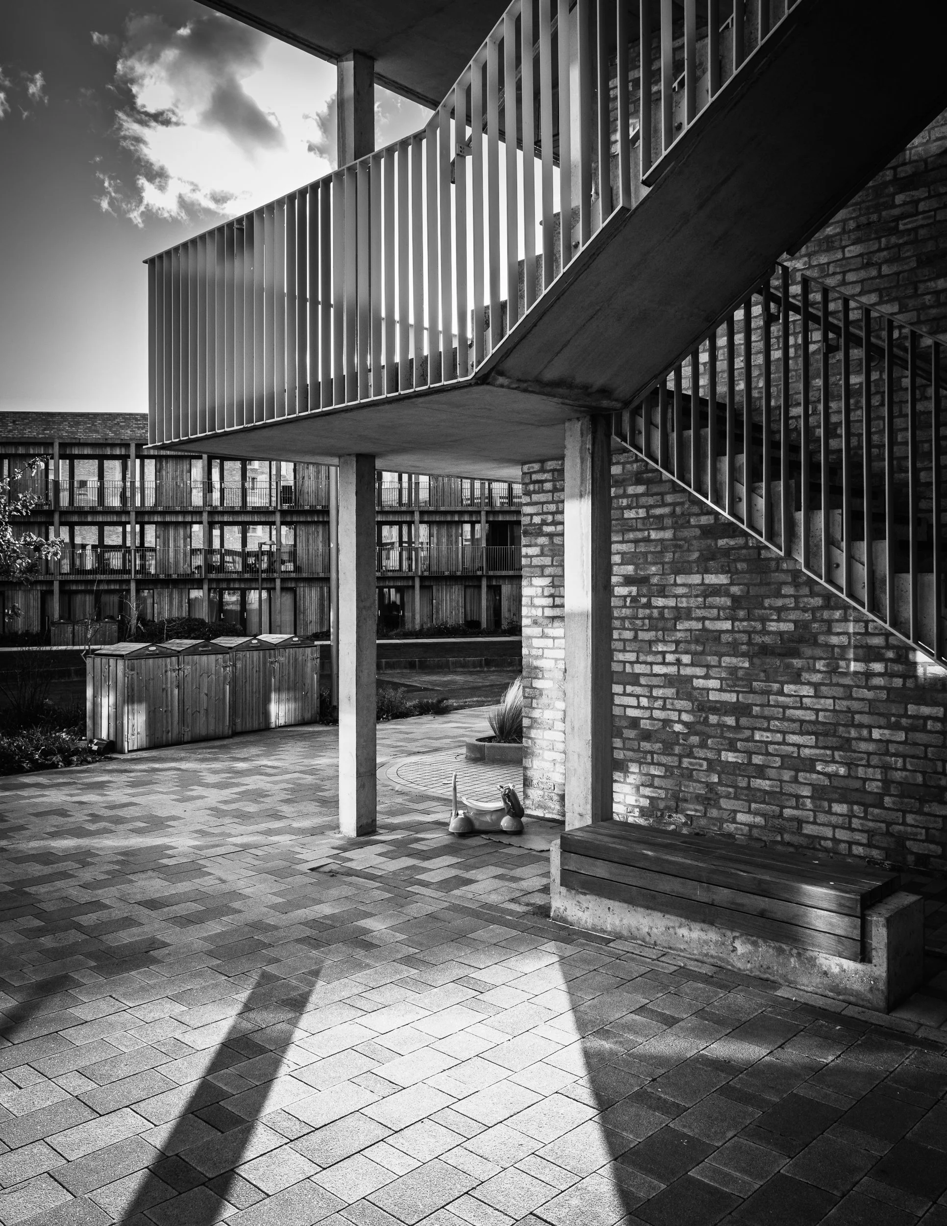 Black and white photo of an outdoor stairway with metal railings, brick walls, and a patio with pavers, shadow play, and some garden elements in the background.
