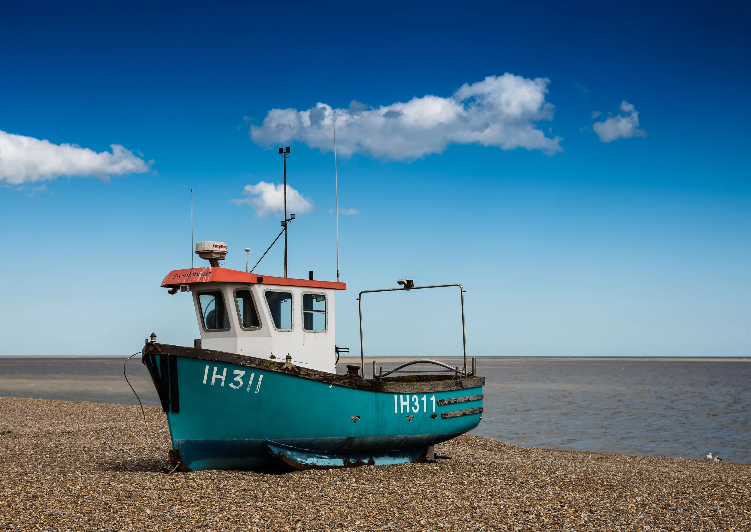 A small blue fishing boat with registration IH311 resting on a pebbled beach under a partly cloudy blue sky, facing towards the sea.