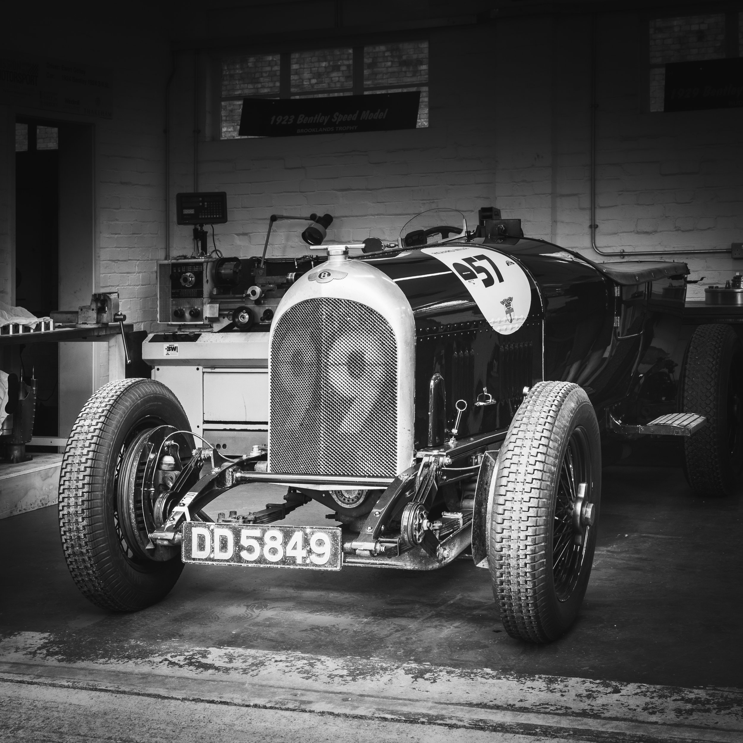 Black and white photo of a vintage racing car inside a garage. The car has a large front grille, exposed wheels, and the number 57 on its side. A license plate reads DD 5849. The garage has tools and equipment on workbenches and a sign on the wall re