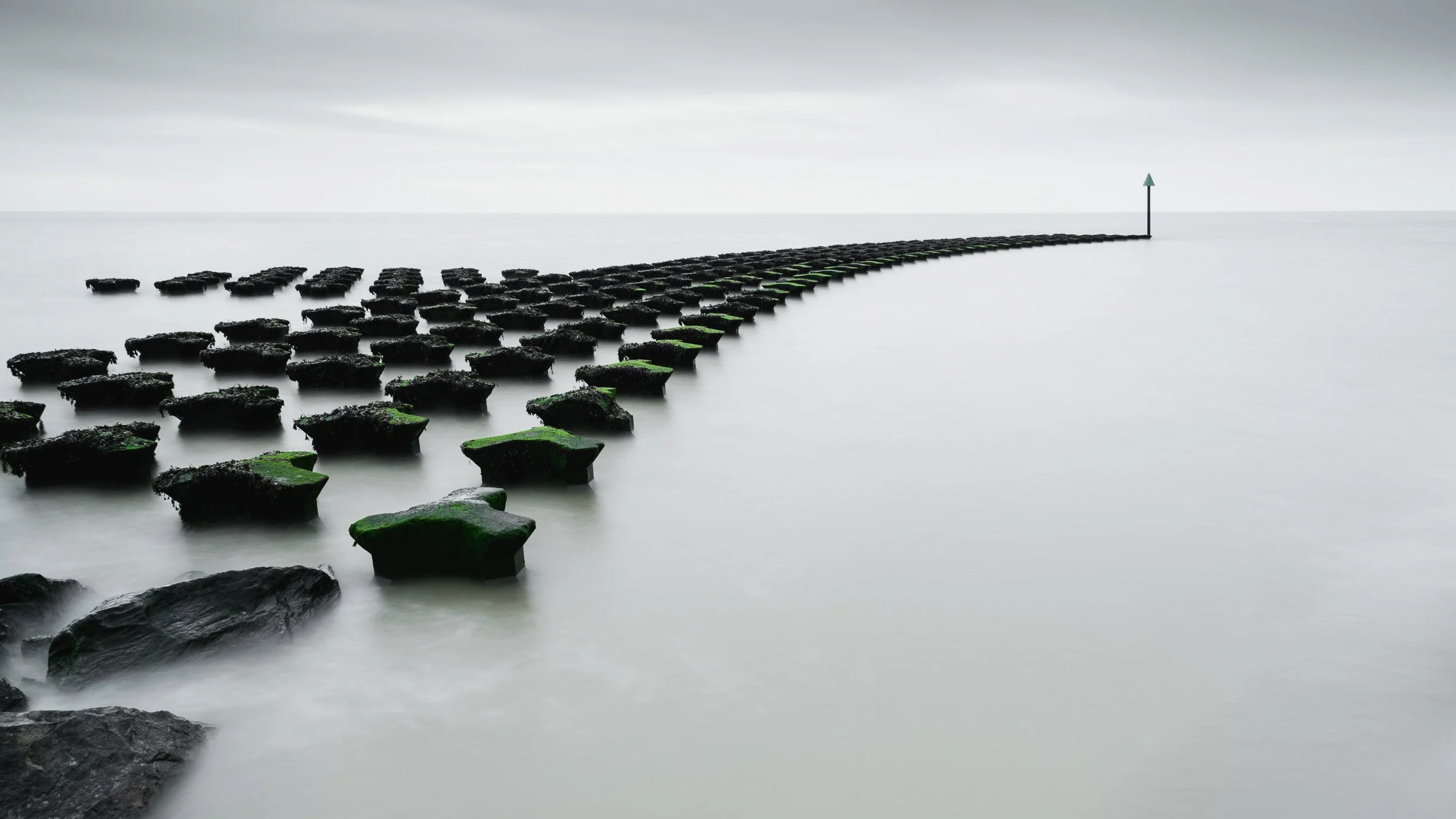 Long row of moss-covered concrete blocks extending into the ocean, forming a pathway toward a signpost with an arrow pointing right, under a cloudy sky.