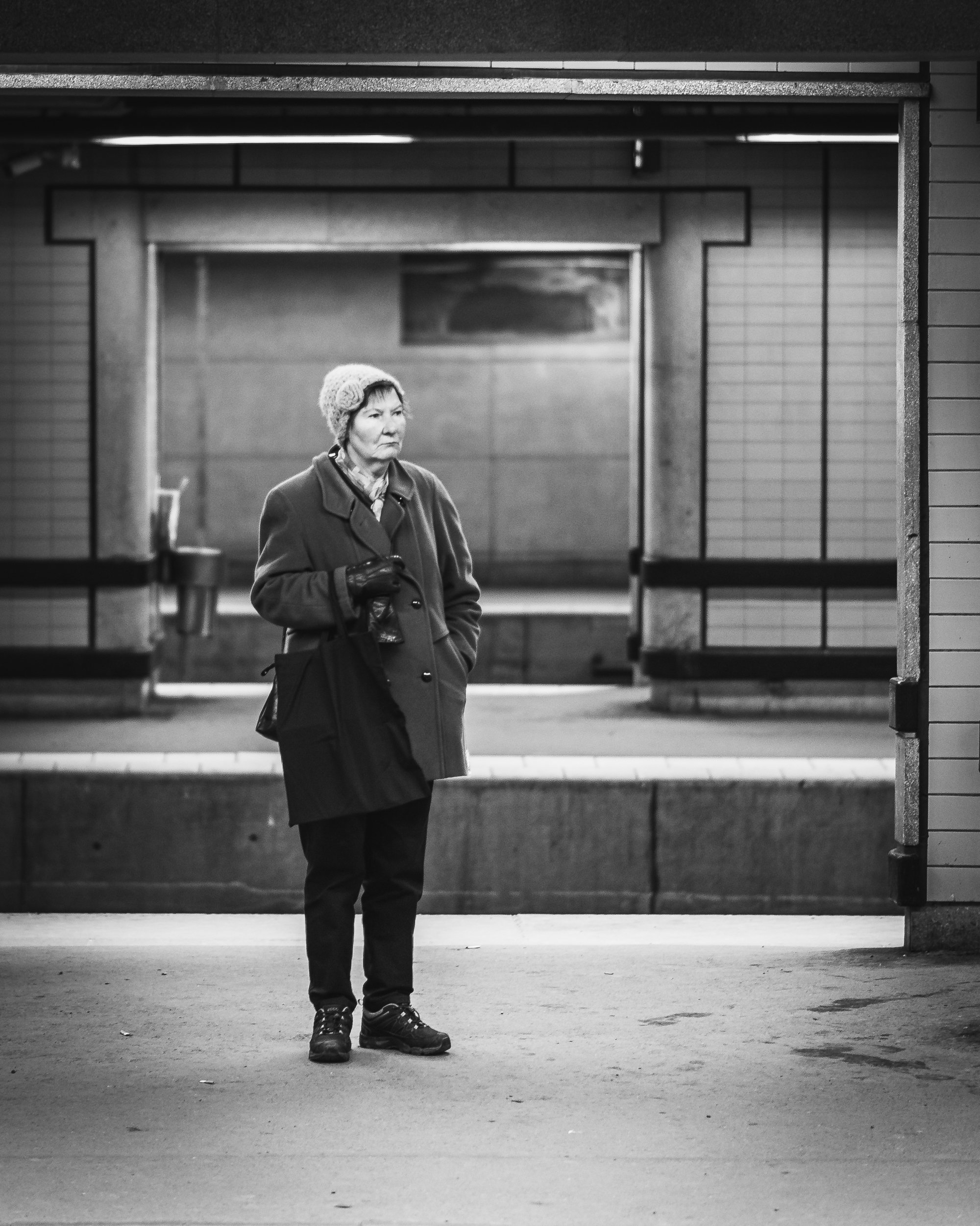 A woman standing alone in an underground metro station, wearing a coat, a hat, and gloves, holding a bag.