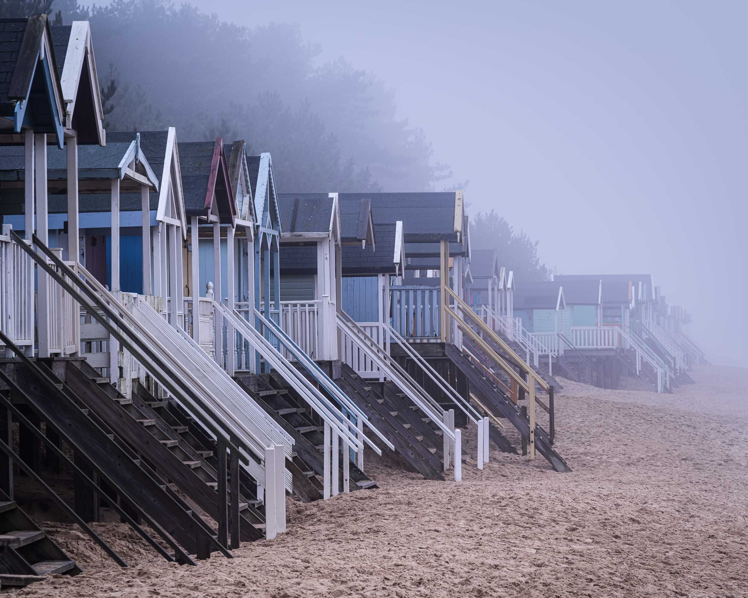A row of colorful beach houses with stairs leading up to their entrances, on a sandy beach with foggy background.