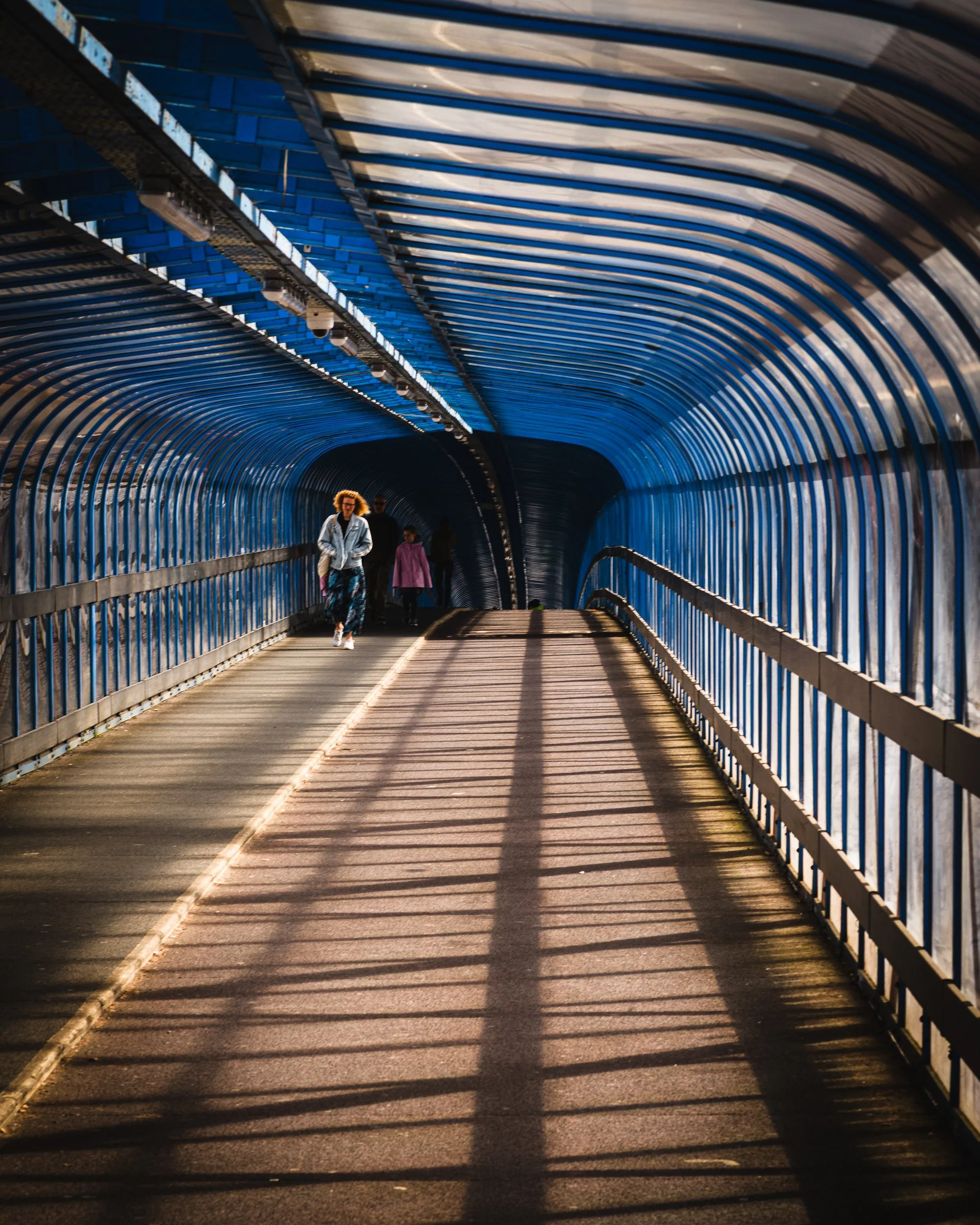 People walking through a blue, curved pedestrian tunnel with shadows cast by the railings on the pathway.