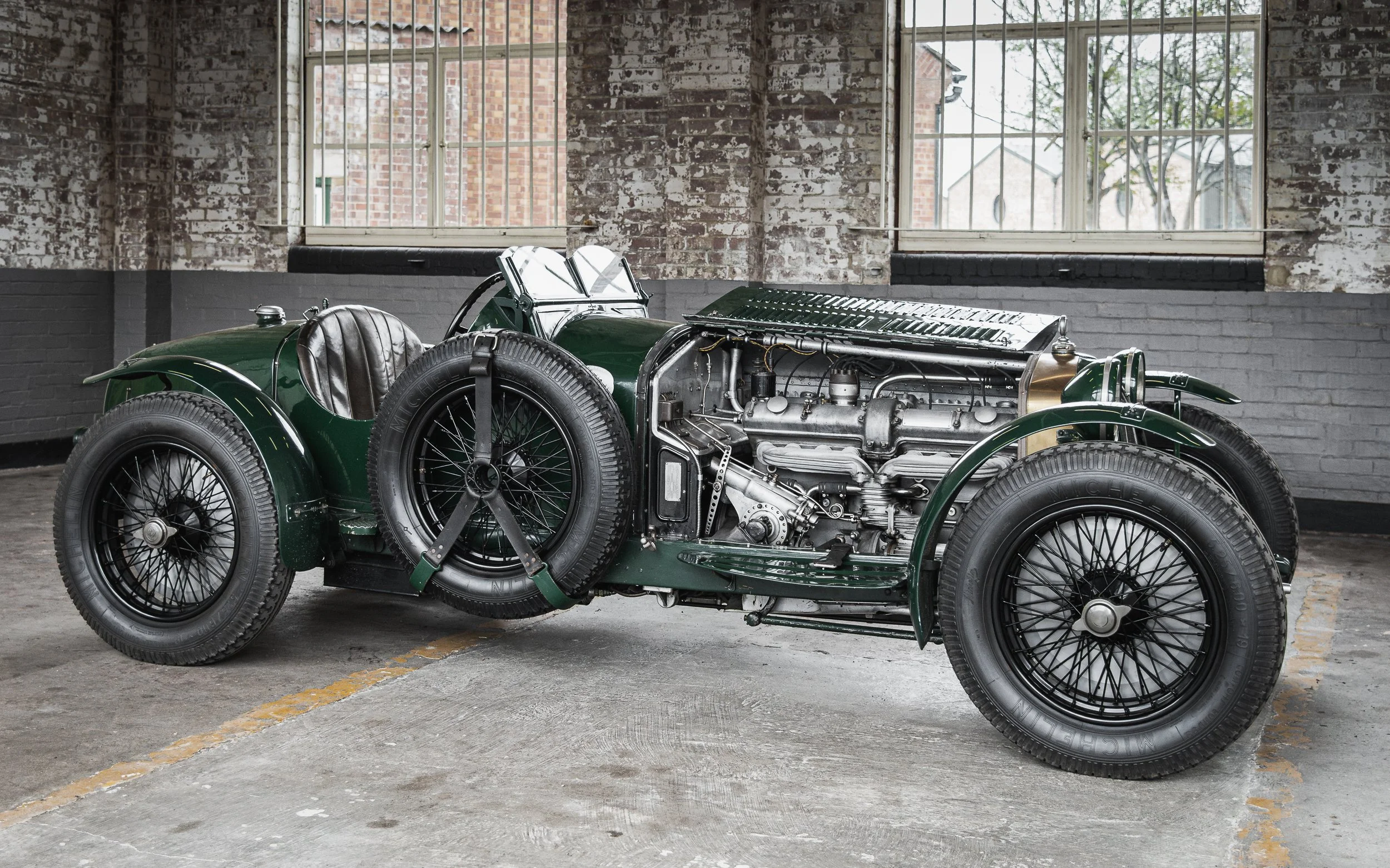 A vintage green racing car with exposed engine parts, large black tires, and a leather seat, parked inside a brick-walled building.