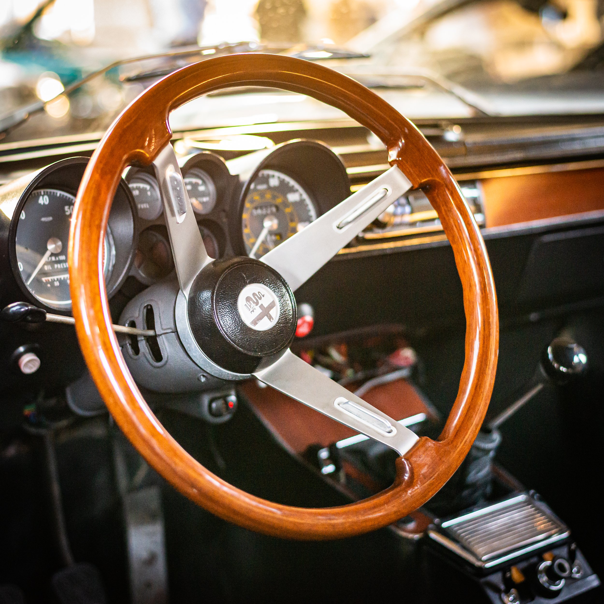 Close-up of a vintage car interior focusing on a wooden steering wheel with an Alfa Romeo emblem at the center, and an dashboard with analog gauges.