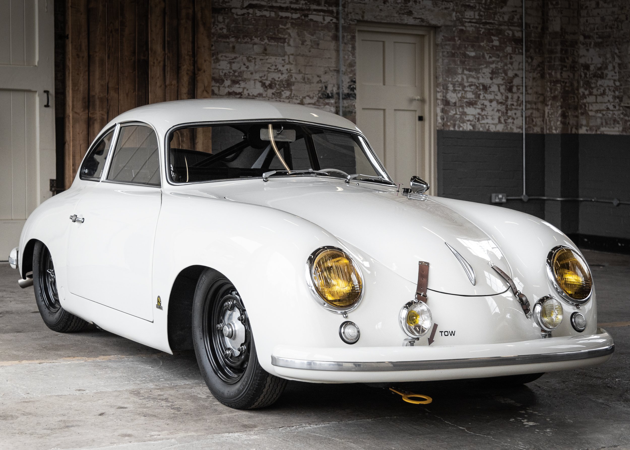 A vintage white race car with yellow headlights parked indoors on a concrete floor, with a rustic brick wall and wooden panels in the background.
