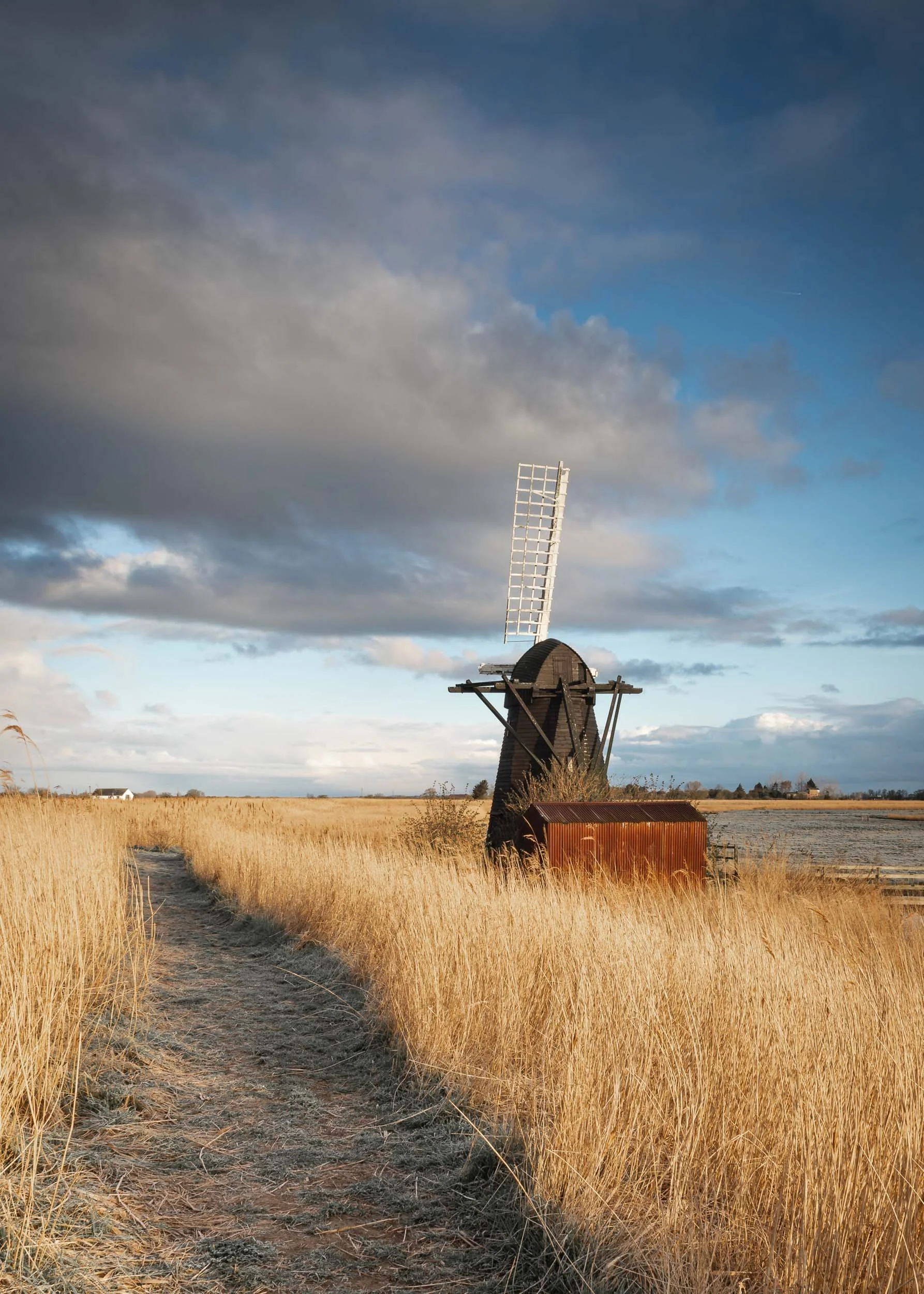 An old windmill standing in a field of tall, dry golden grass with a dirt path leading towards it, under a cloudy sky with patches of blue.