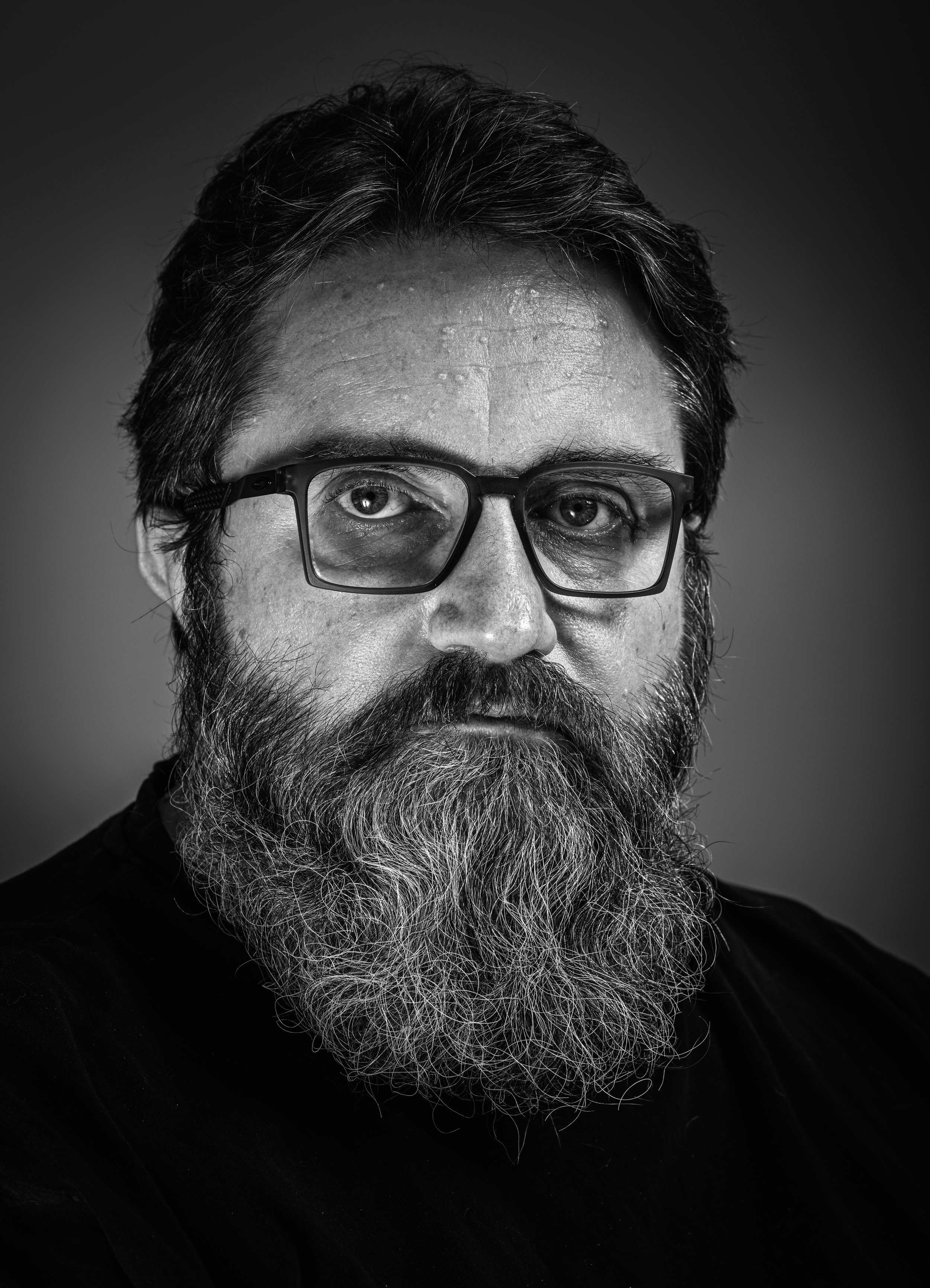Black and white portrait of a man with glasses, a full beard and mustache, looking directly at the camera with a serious expression.