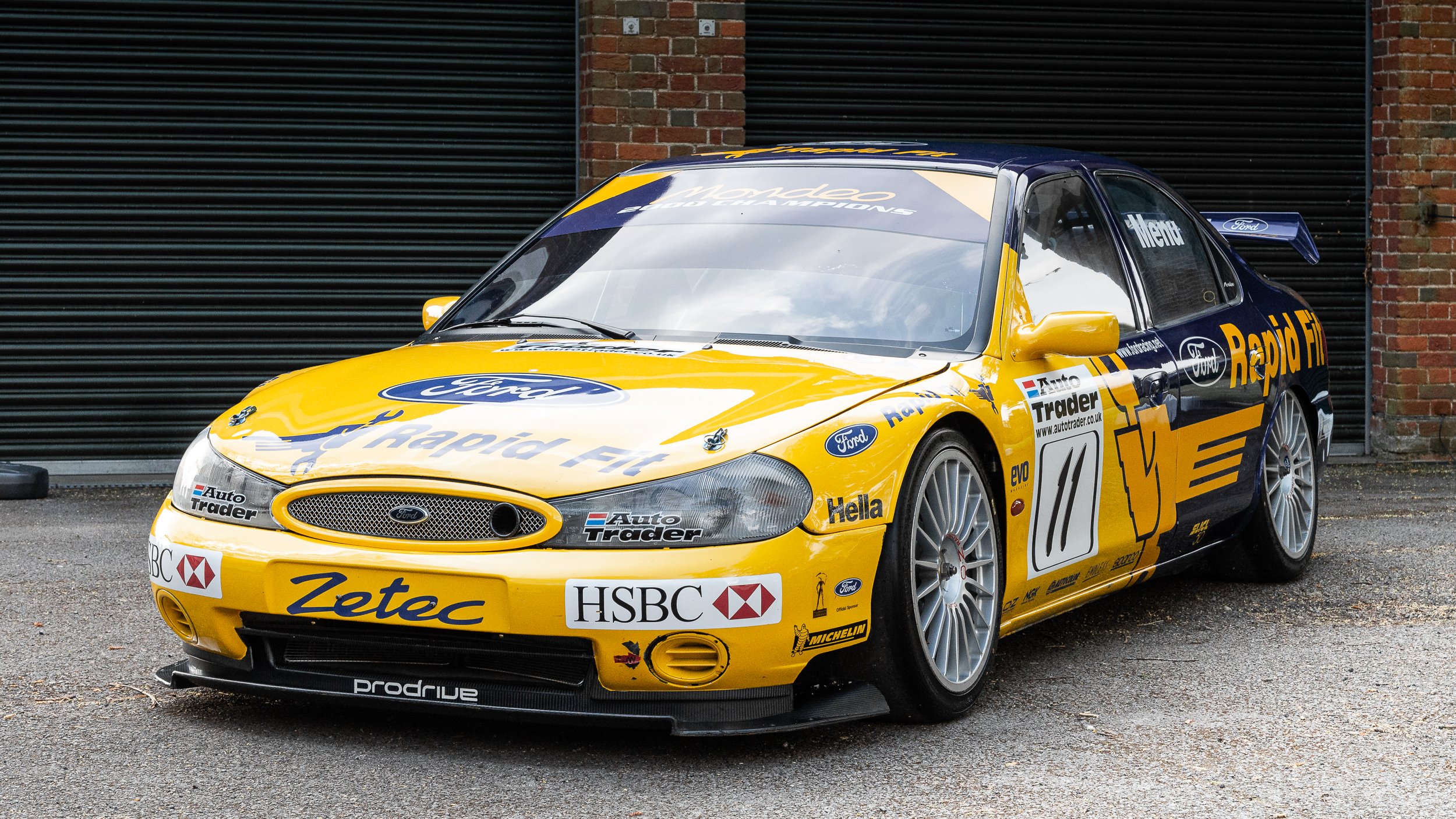 Yellow and blue Ford race car with sponsor logos, parked on asphalt in front of a black roller door.