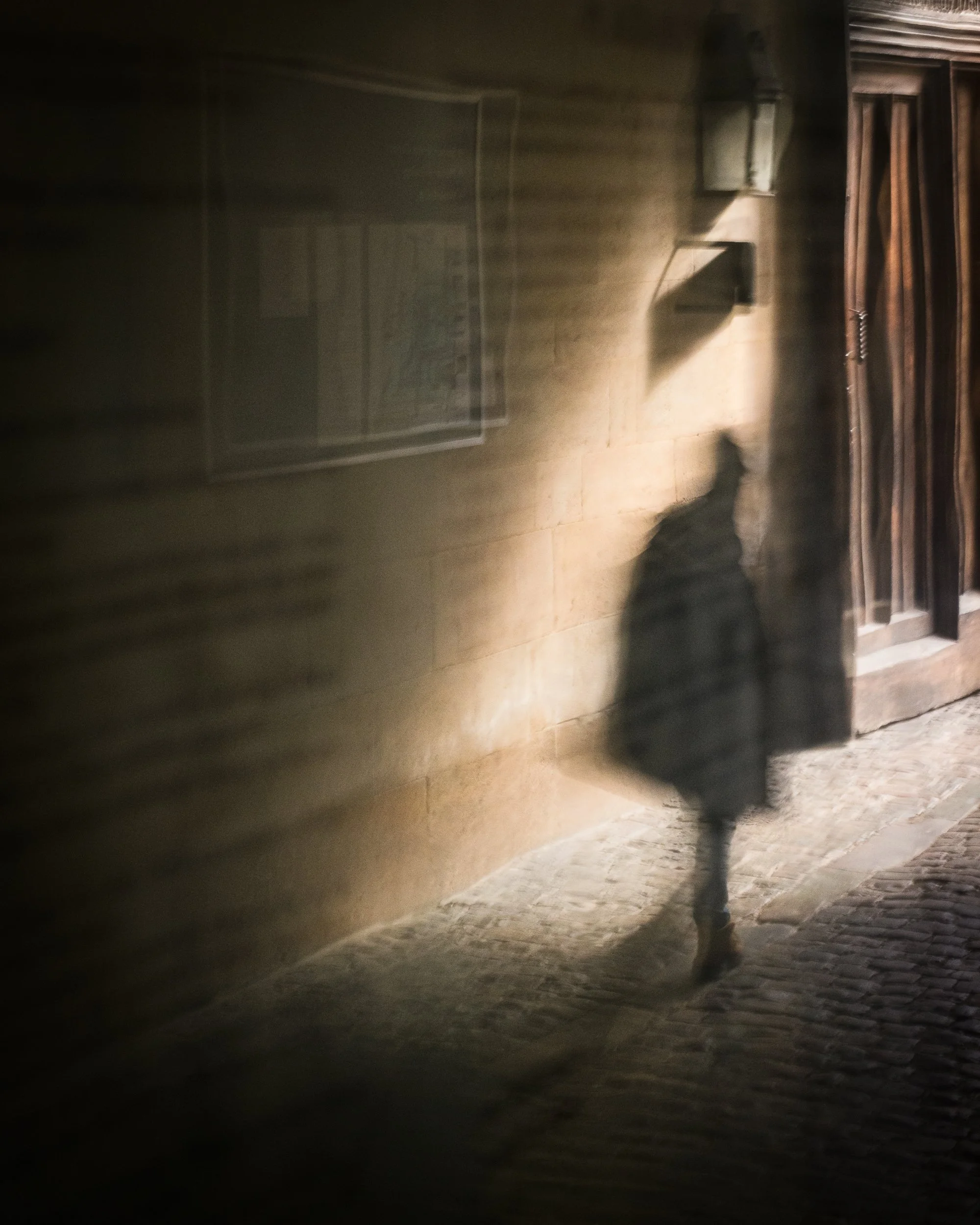 A shadow of a woman walking on a cobblestone street at night, with a beige brick wall and wooden door in the background, illuminated by a streetlamp.