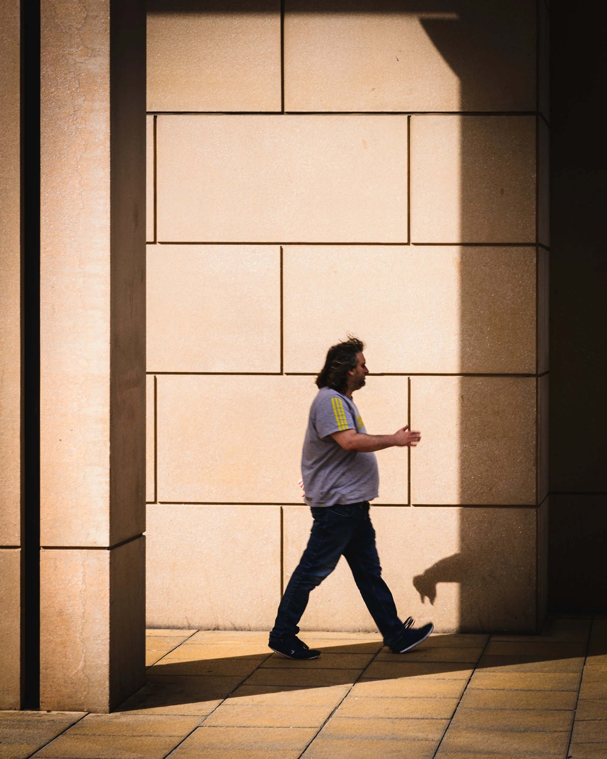 A man with long hair walking past a stone wall, casting a shadow on it, in daylight.