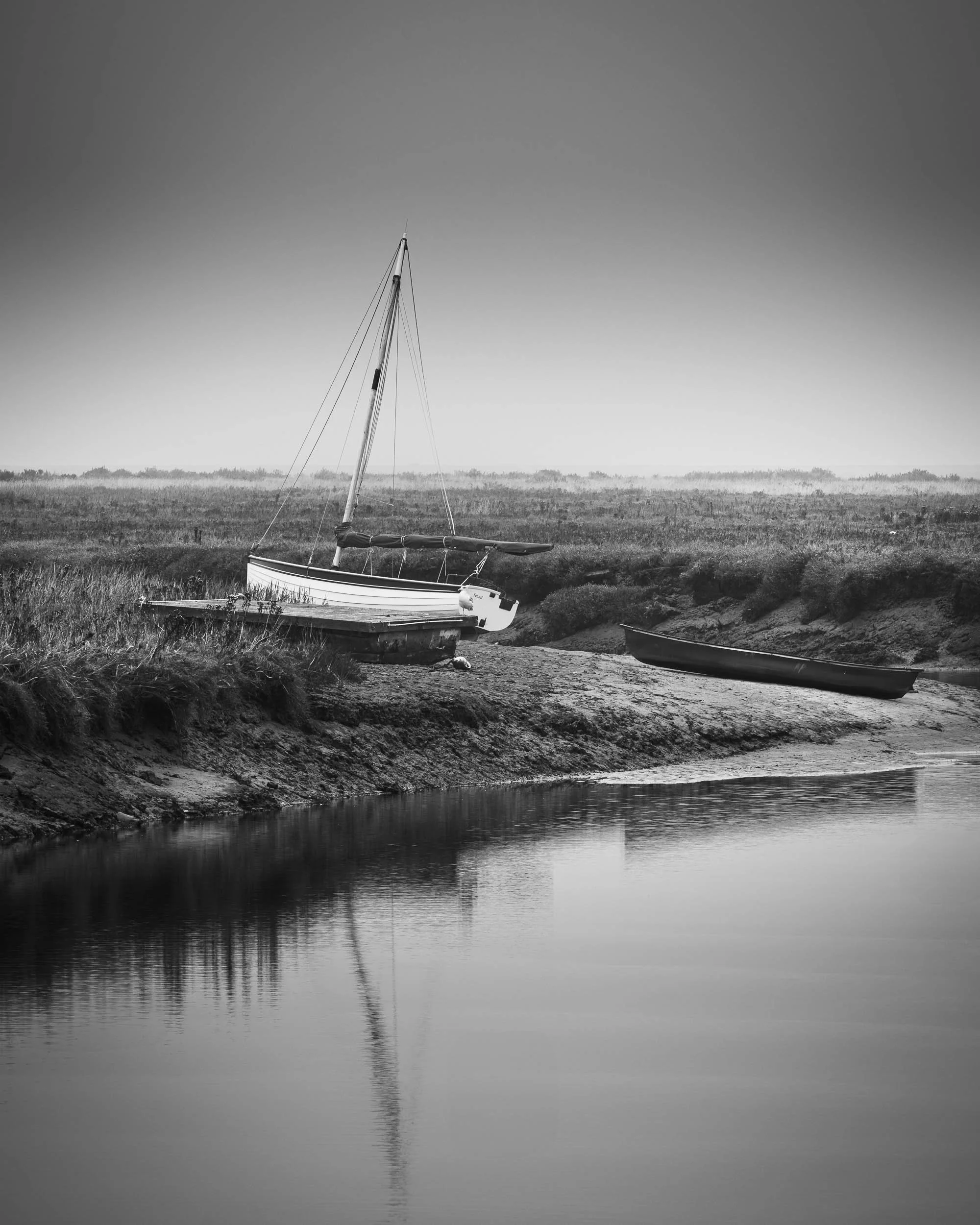 Black and white photo of a sailboat on a small shore with a calm body of water reflecting the boat, and a small rowboat nearby in a natural landscape.
