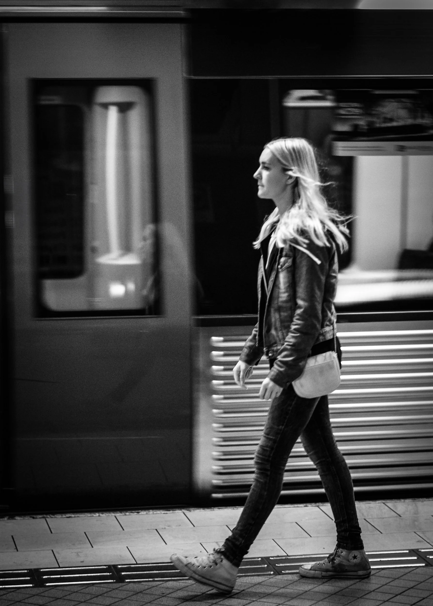 A young woman with long hair walking past a train or subway in an underground station. She is wearing a leather jacket, jeans, and white sneakers, and carries a small shoulder bag.