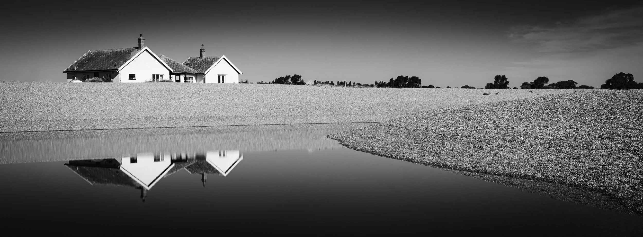 Black and white photo of a house with chimneys located near a body of water, with trees and bushes in the background and reflections in the water.