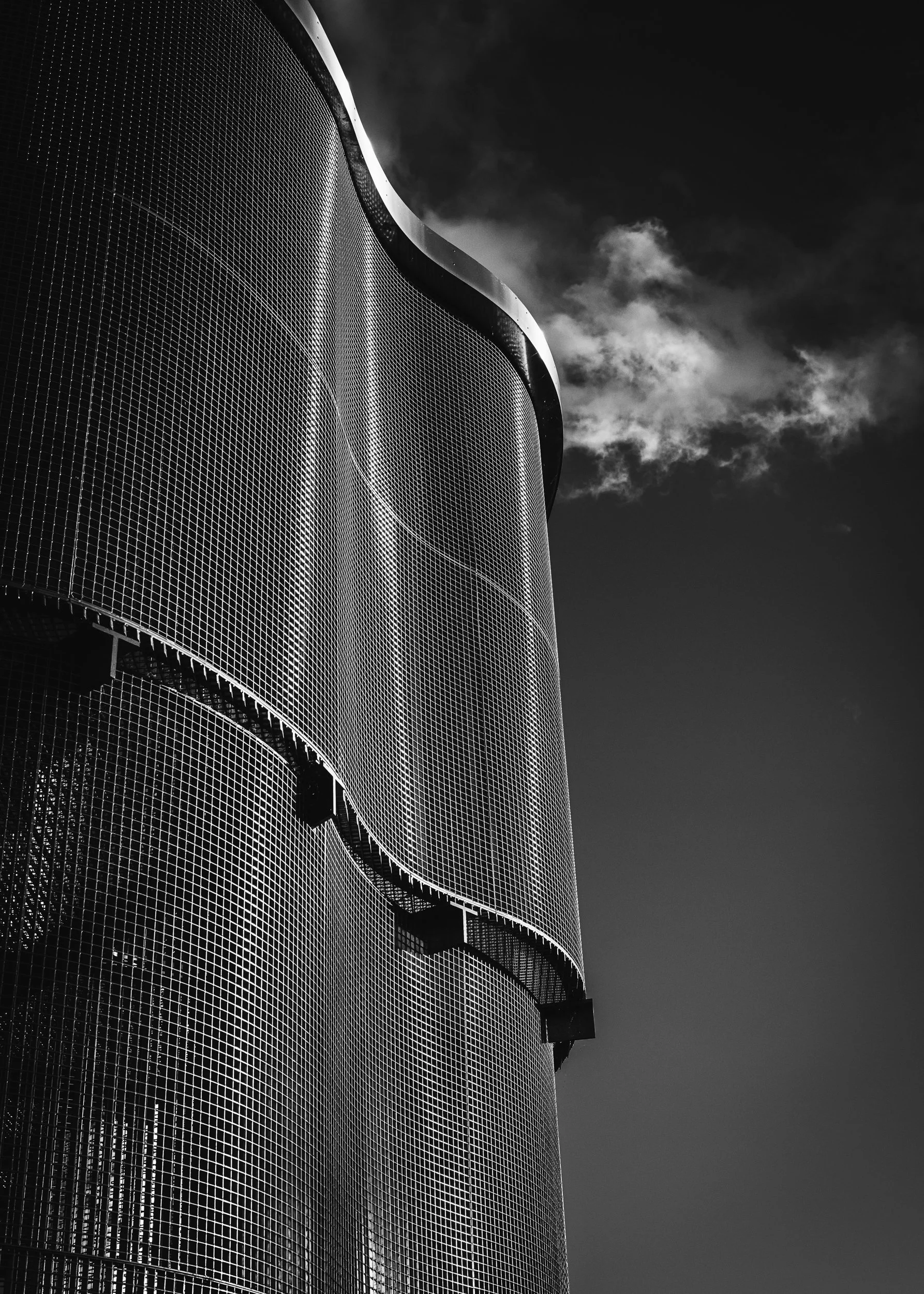 A modern cylindrical building with a textured facade and curved design, set against a cloudy sky in black and white.