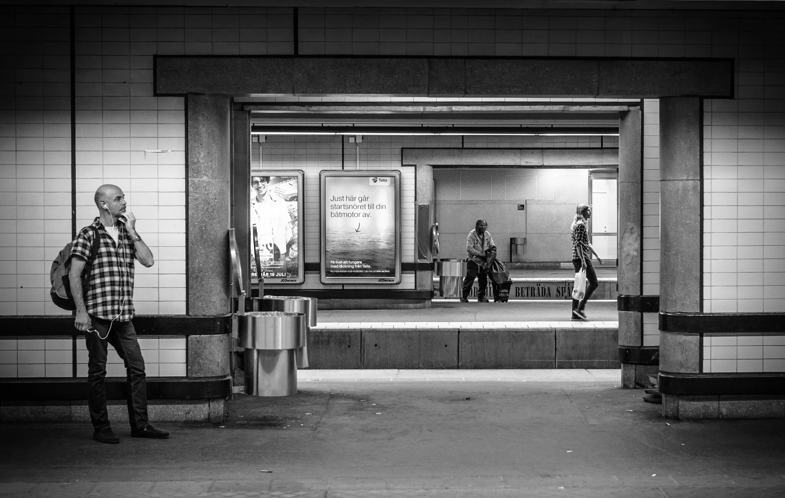People waiting at an underground subway station, some with luggage, in black and white.
