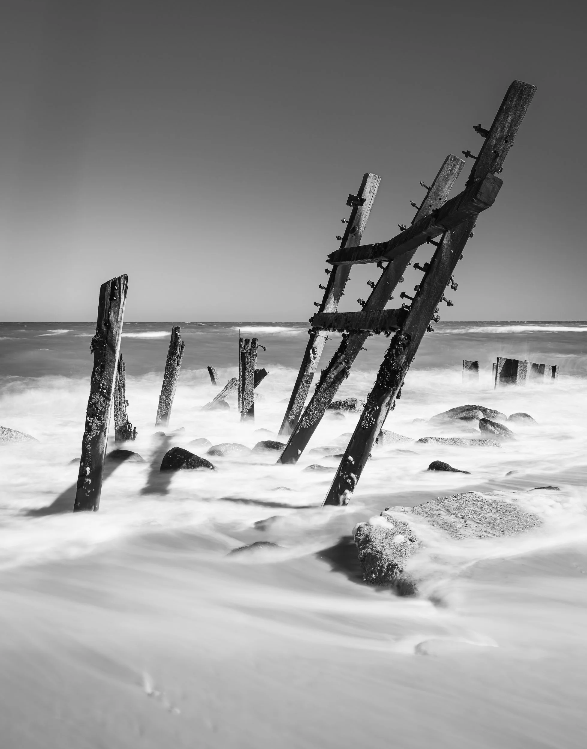 Black and white photo of weathered, broken boat remains along a shoreline, with waves washing over the rocks and sand.