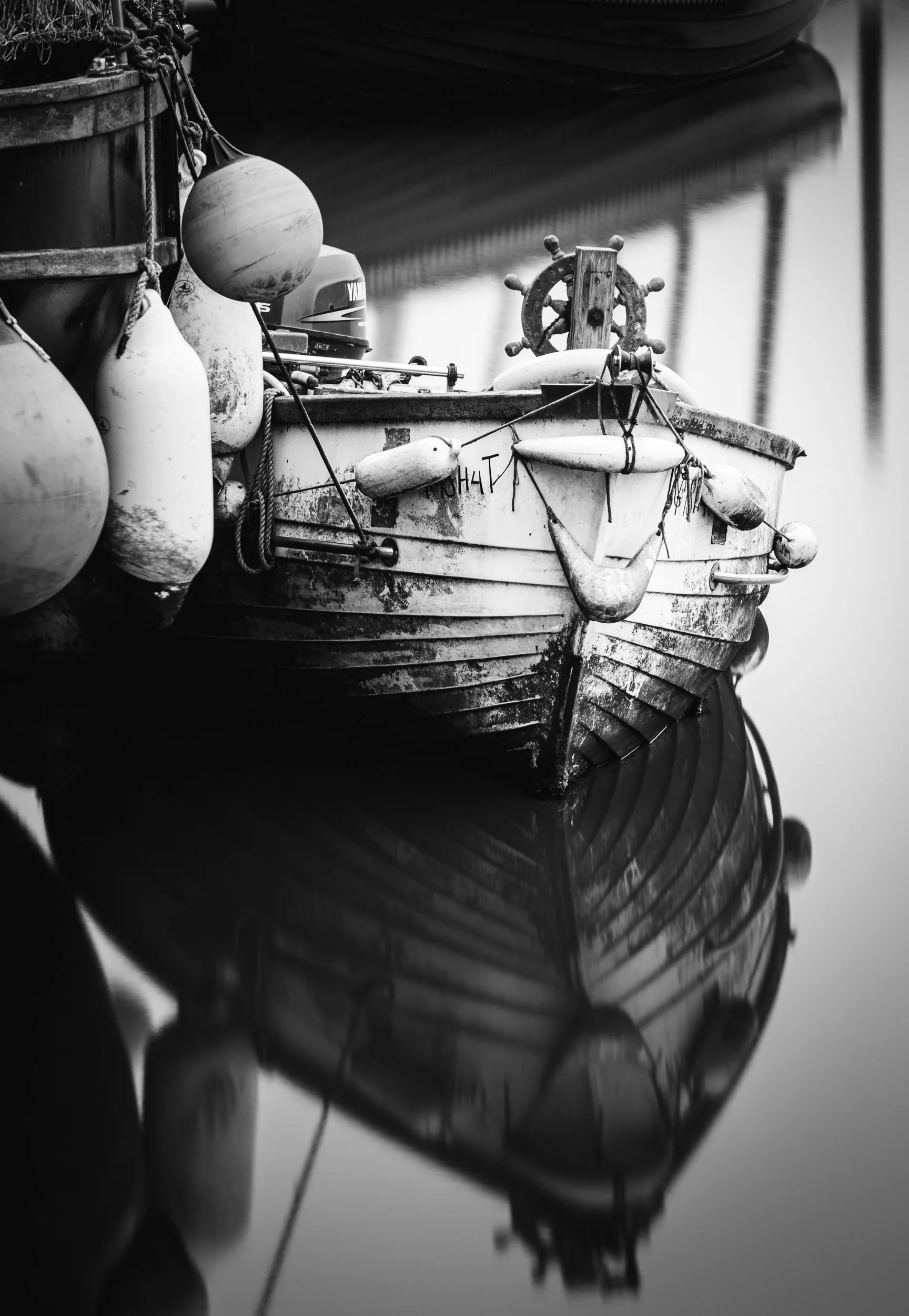 A black and white photo of a small, weathered boat docked in calm water, with numerous fenders hanging from its side and a steering wheel at the bow. The boat's reflection is visible in the water.