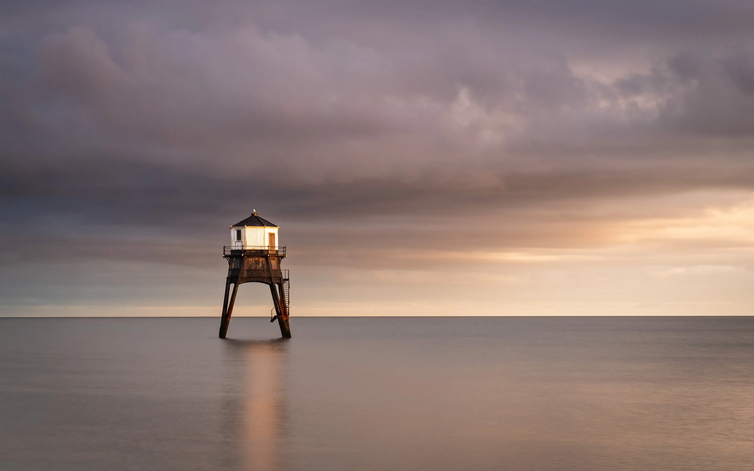 A solitary lighthouse standing on stilts in calm water, with a cloudy sky during sunset.